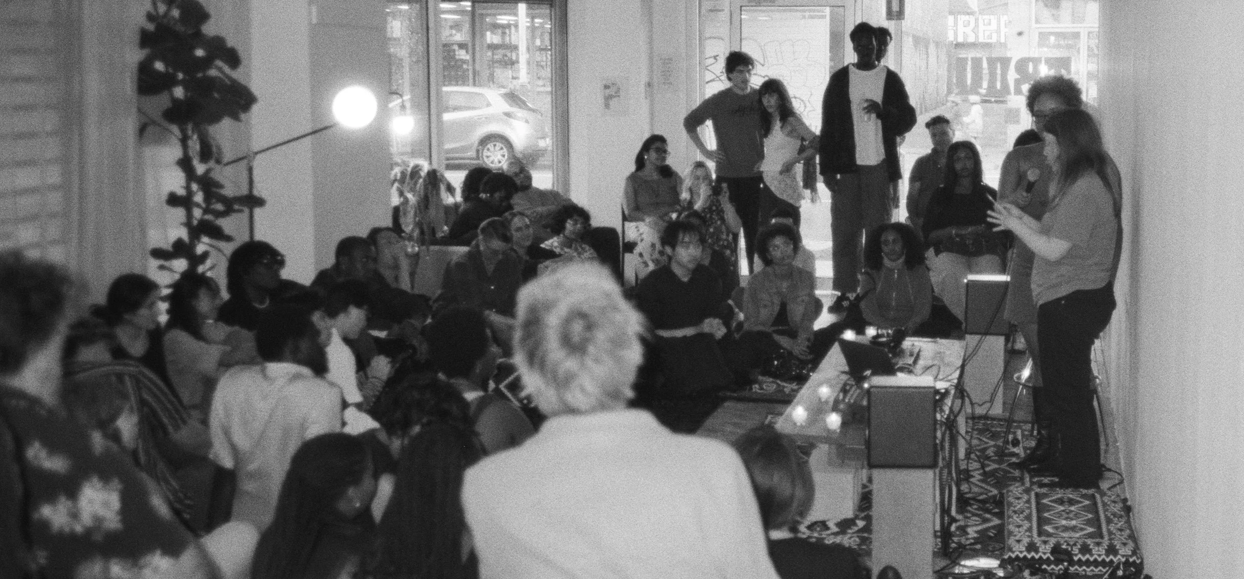 A group of people gathered indoors for a presentation or event, with a woman speaking at the front and the audience attentively listening.