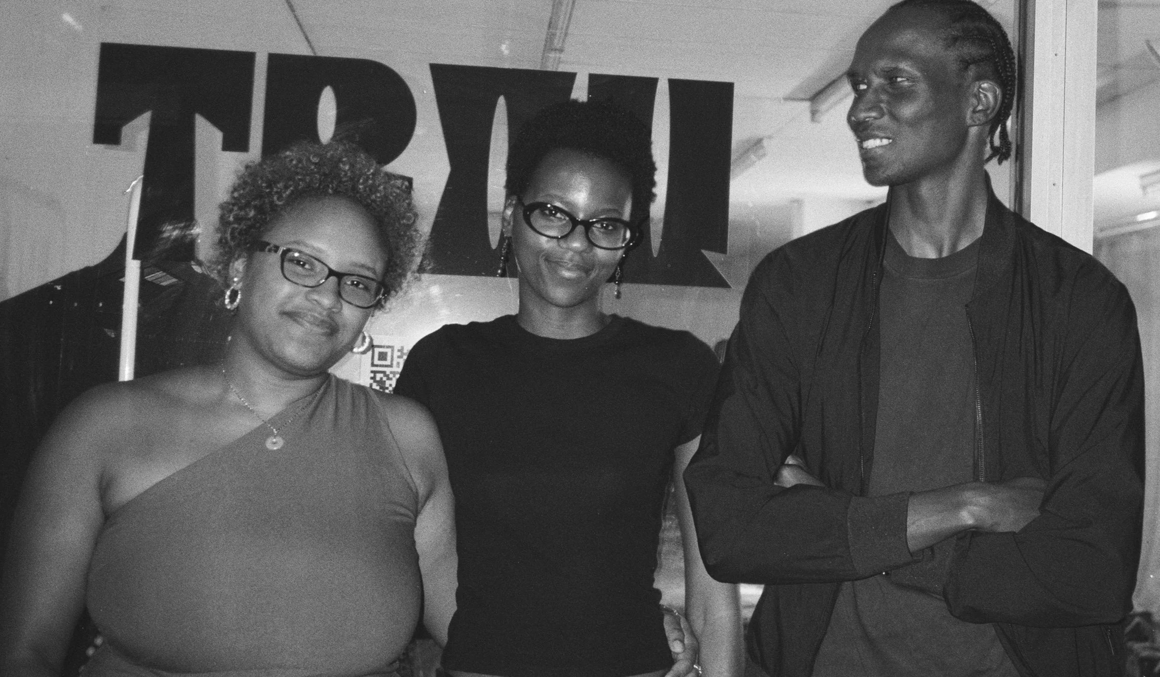 Three women and one man are standing together indoors. They are dressed nicely, with the women wearing glasses and earrings, and the man wearing a jacket. The woman on the right is shaking hands with the man. There is a sign behind them that says 'TR