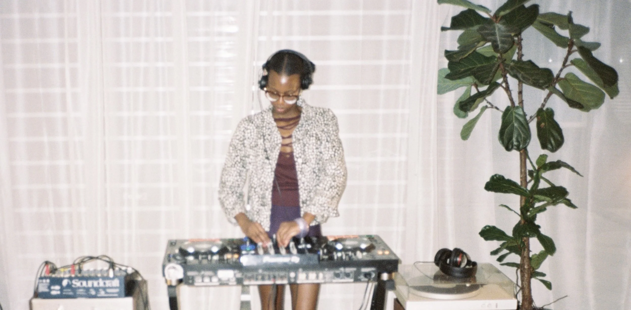 A woman with curly hair and glasses, wearing a patterned jacket and maroon shirt, is DJing with turntables and audio equipment on a table in front of a white curtain. There is a potted plant with large, green leaves to her right, and headphones resti