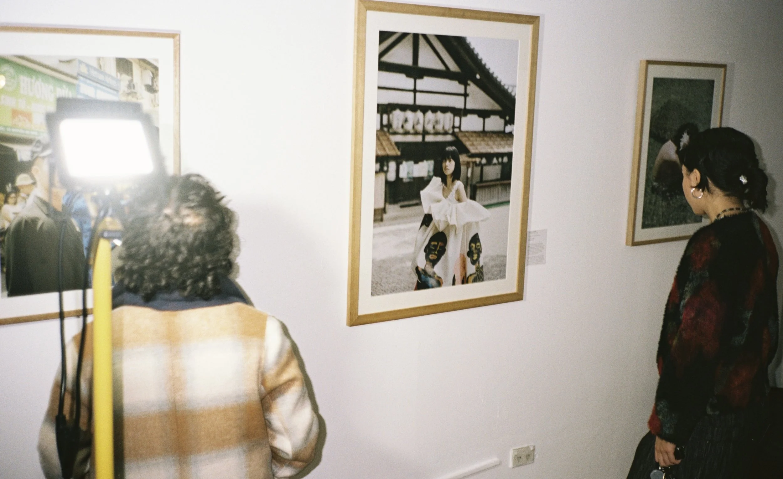 People viewing artwork in an art gallery, with framed pictures on the wall, including a woman in a white dress at a train station, and another woman in a dark top with earrings, in the foreground.