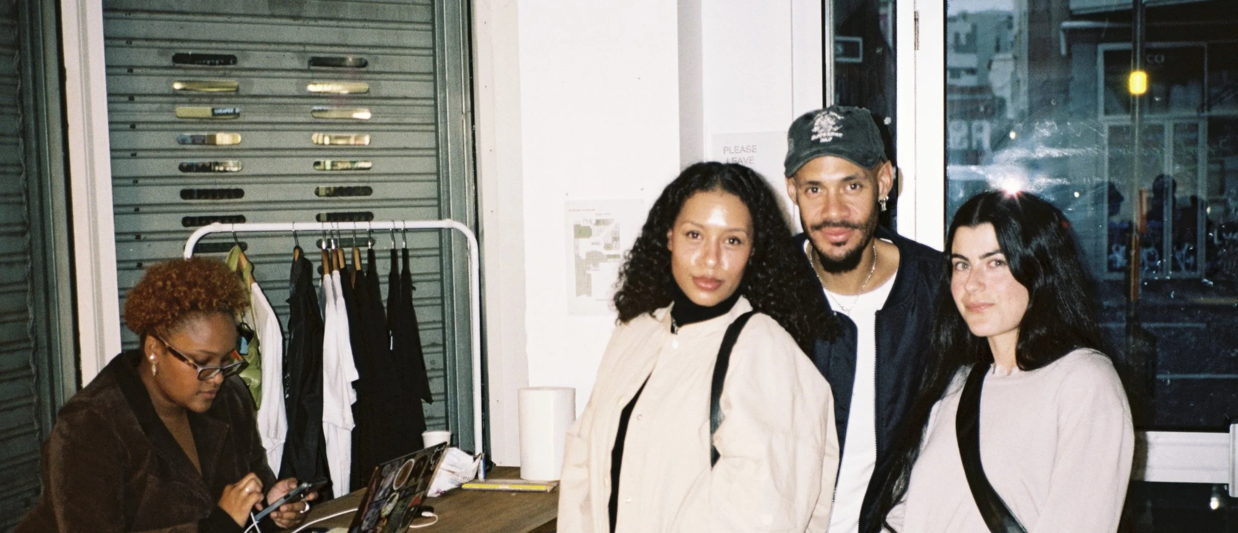 Four people standing inside a store, with a woman sitting at a table and looking at her phone. There are clothes hanging on a rack beside her and a window behind the group.