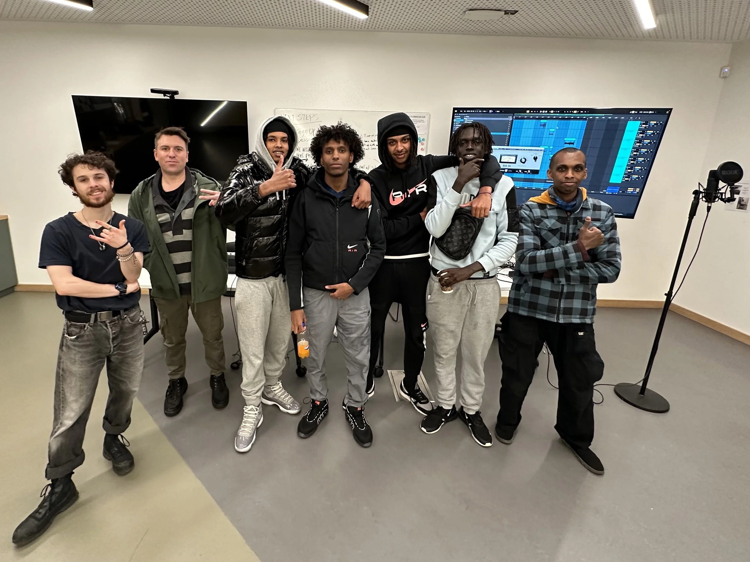 Group of seven young men standing together in a room with electronic music equipment and screens in the background.