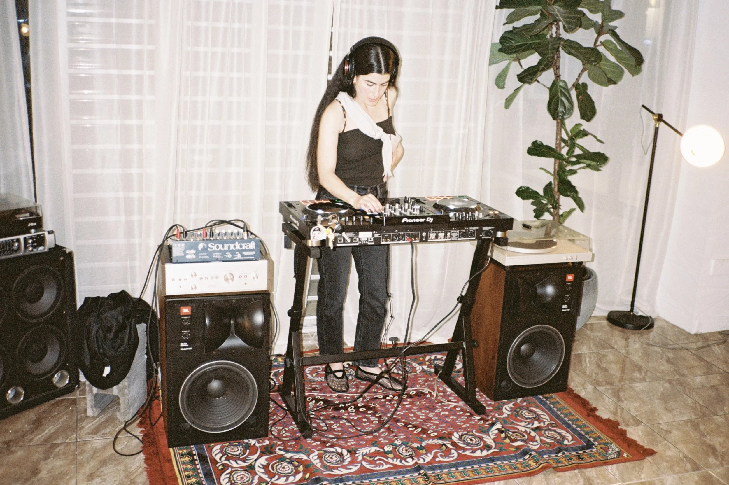 A woman with long dark hair and light skin DJing in a room with a white curtain, a large plant, and speakers, standing at a DJ console on a small table on a colorful rug.