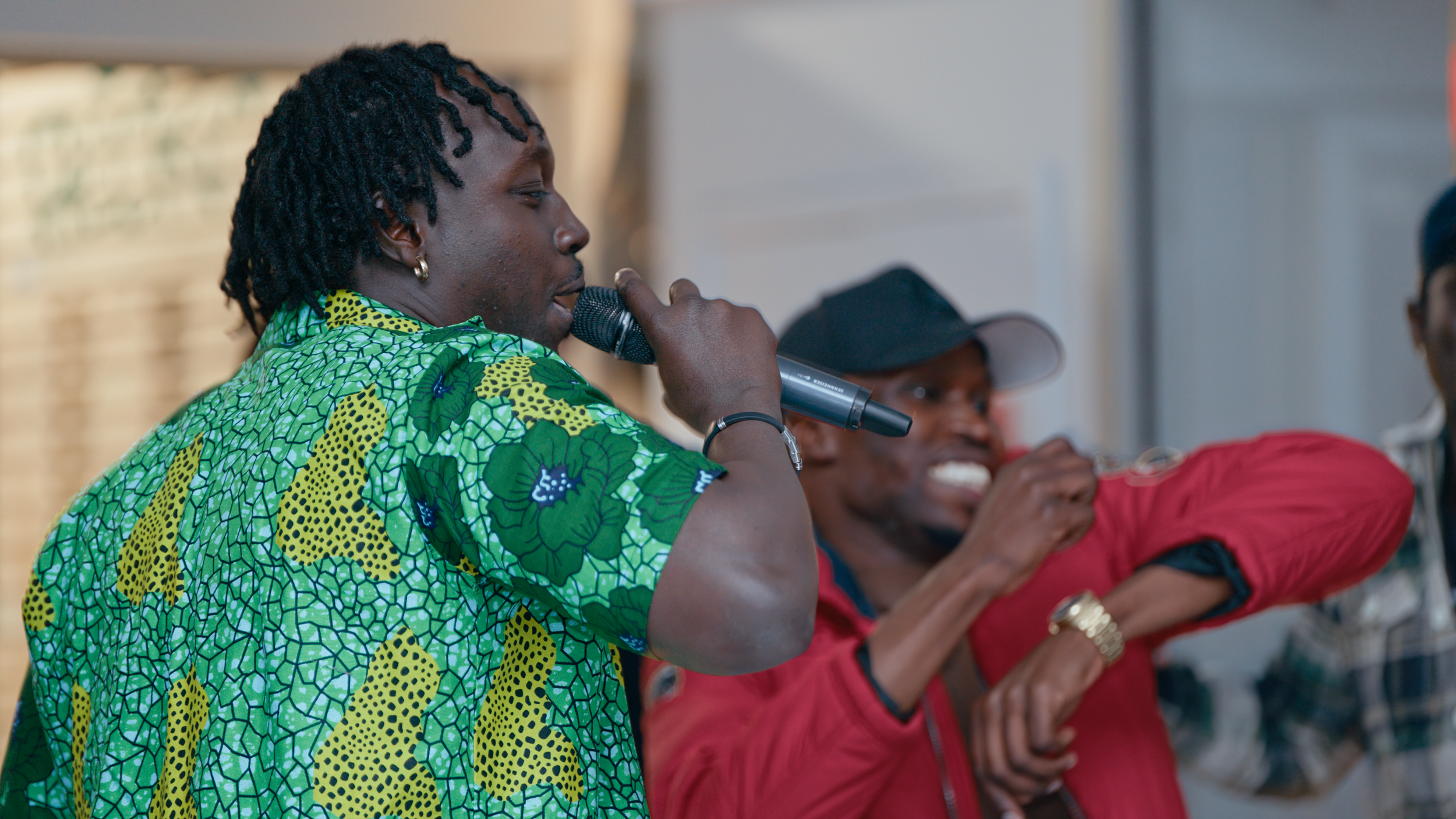 A person wearing a green patterned shirt speaking into a microphone, with two people sitting next to them, one in red and one in black, smiling and holding a utensil.
