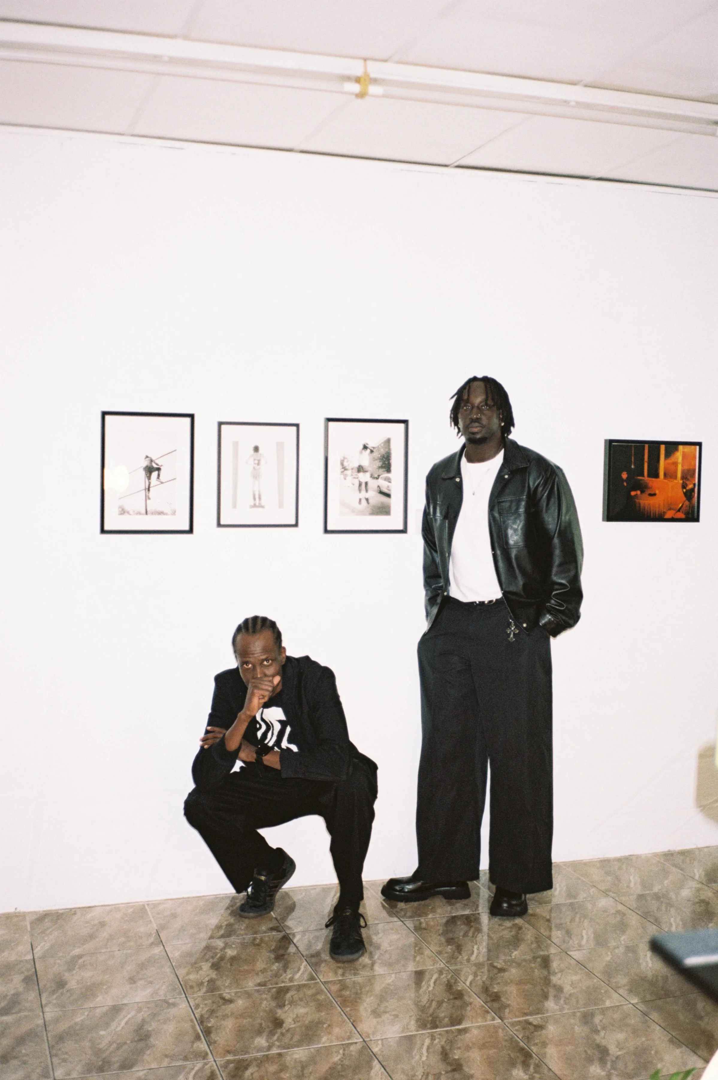 Two men pose in front of framed photographs on a white gallery wall. One man is crouched, resting his chin on his hand, while the other stands with hands in pockets. Both wear dark clothing, and the space has tiled floors.