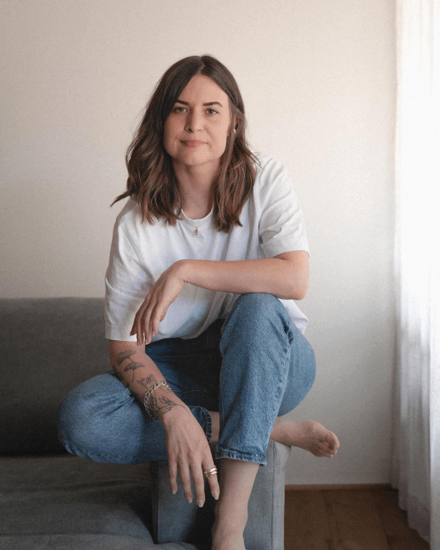 Junge Frau mit dunklen, schulterlangen Haaren sitzt auf einer Couch, trägt ein weißes T-Shirt und blaue Jeans, hat Tattoos am linken Arm, einen Ring an der rechten Hand und sitzt mit einem Bein auf dem Boden, das andere Bein gekreuzt.