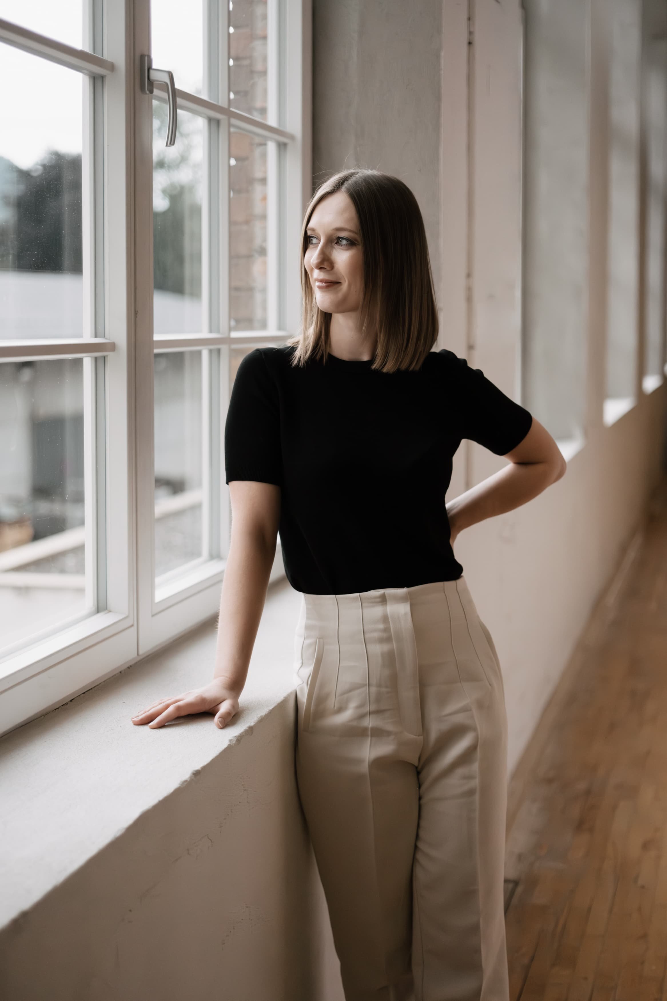 Junge Frau mit braunen Haaren, schwarzem T-Shirt und weißen Hosen, die an einem Fenster im Innenraum steht, mit Blick nach draußen.