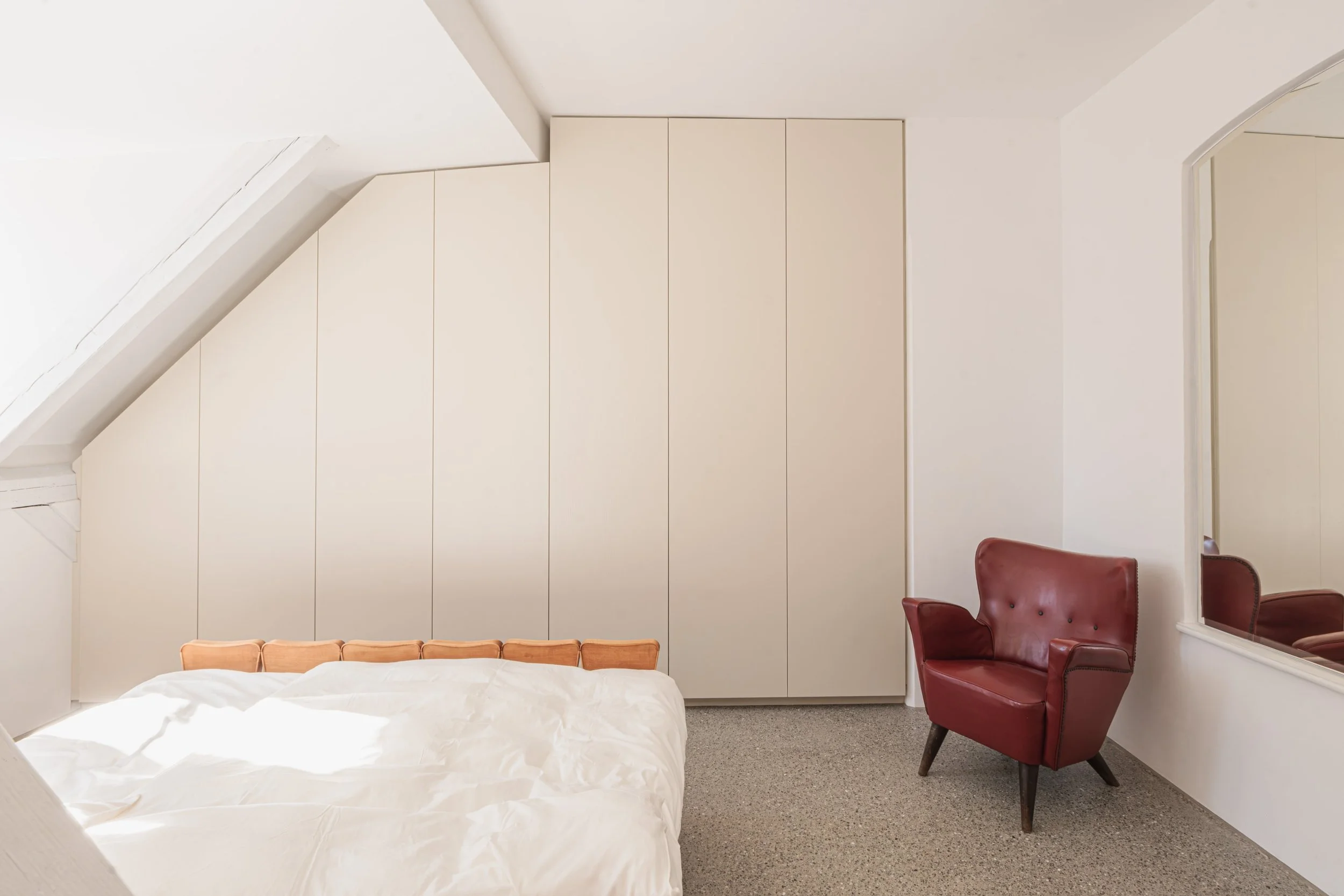 Ein Schlafzimmer mit weißen Wänden, einem Bett mit weißer Bettwäsche, einem roten Ledersessel und einem großen weißen Schrank.