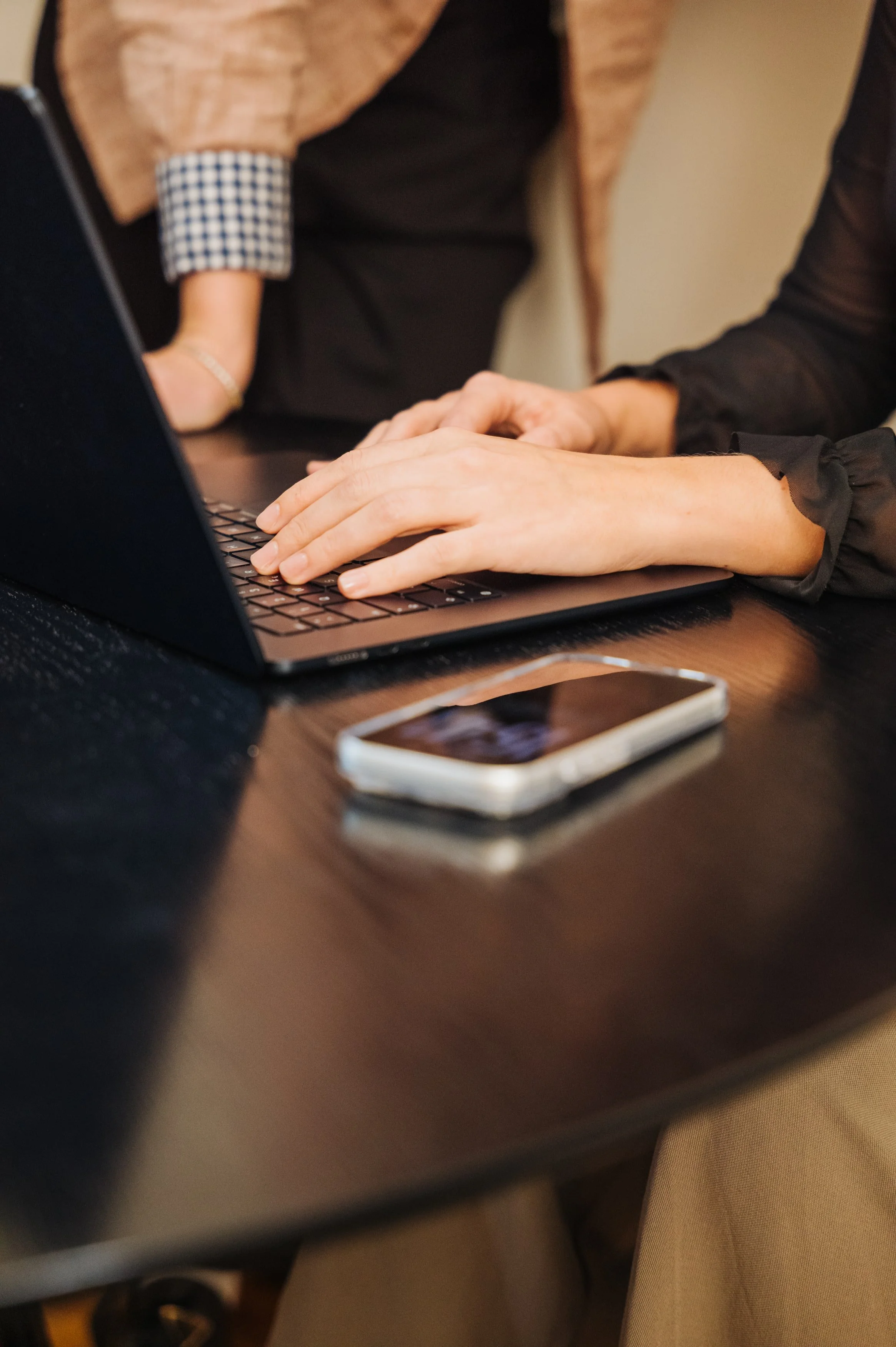Person tippt auf einer Laptop-Tastatur, Smartphone liegt auf einem dunklen Tisch.