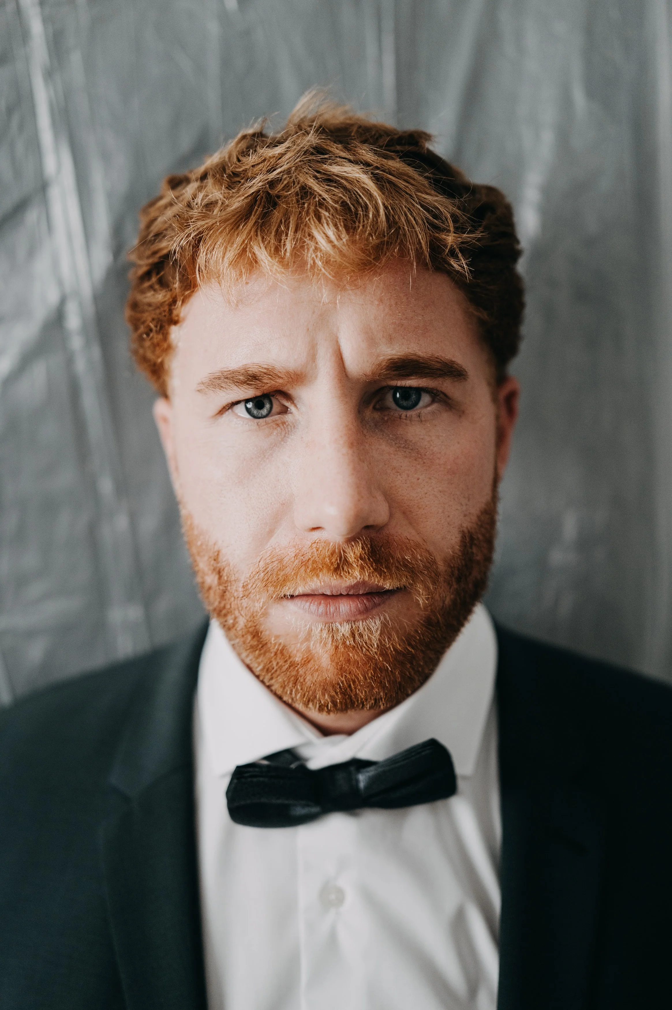 Porträt eines Mannes mit roten Haaren und Bart, formeller schwarzer Anzug, weiße Hemd und schwarzer Fliege, vor einer grauen Wand.
