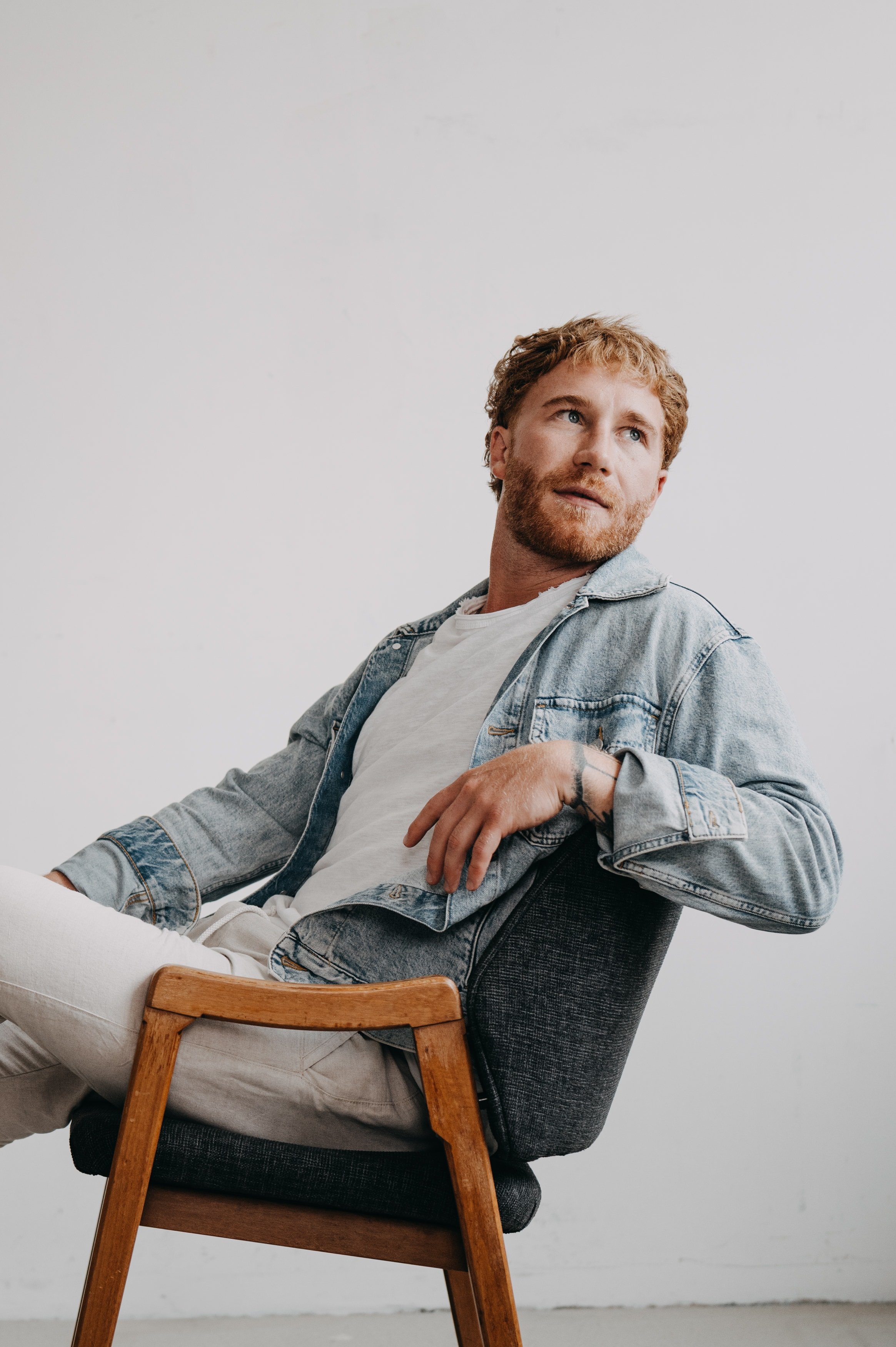 Ein jugendlicher Mann mit roten Haaren, Bart und blauen Augen sitzt entspannt in einem Stuhl, trägt ein weißes T-Shirt und eine denim Jacke, vor einem neutralen Hintergrund.