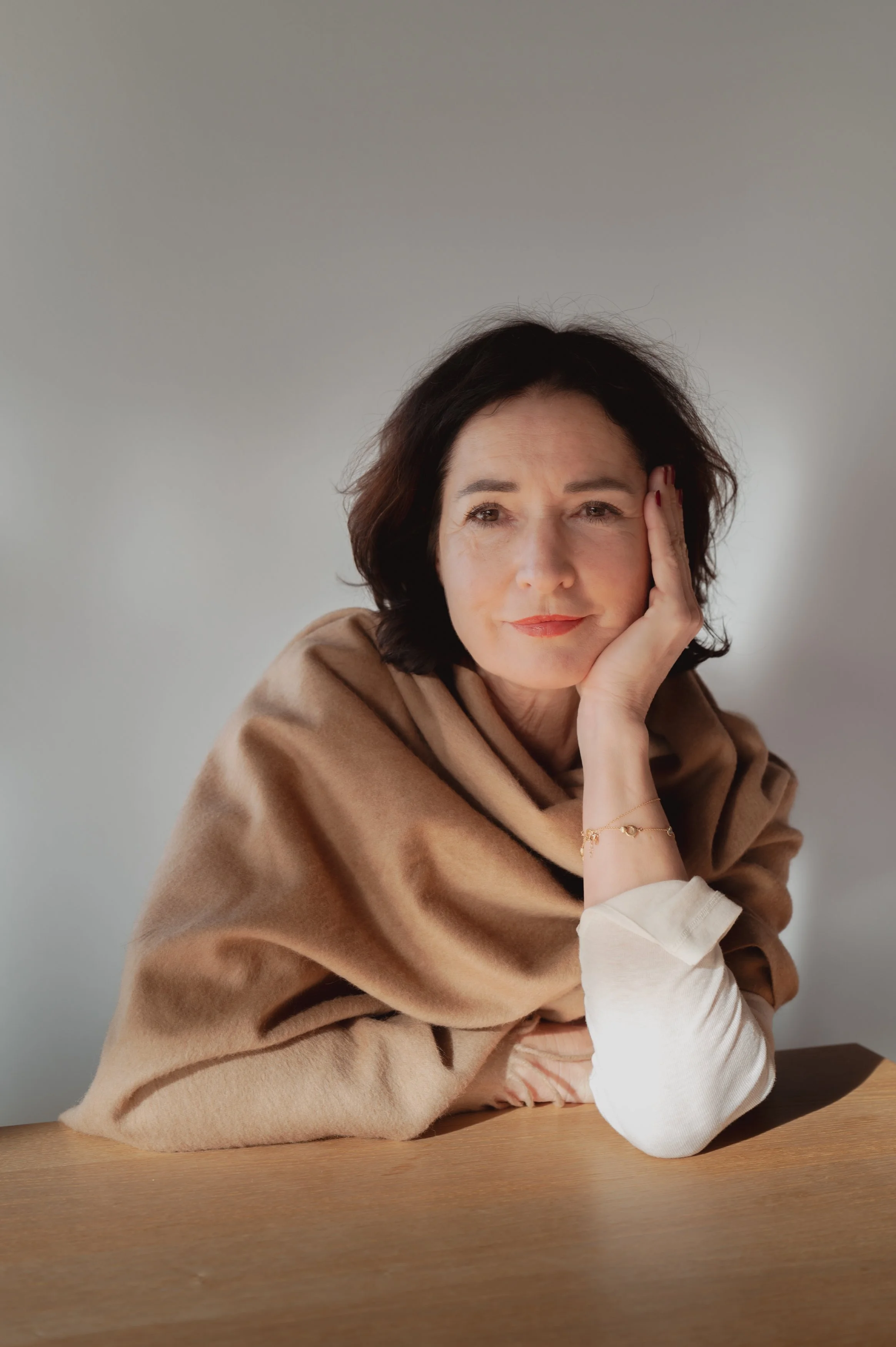 Frau mit dunklem Haar, trägt eine beige Schals und weiße Oberteil, sitzt an einem Holztisch, hat einen nachdenklichen Blick, vor neutralem grauem Hintergrund.