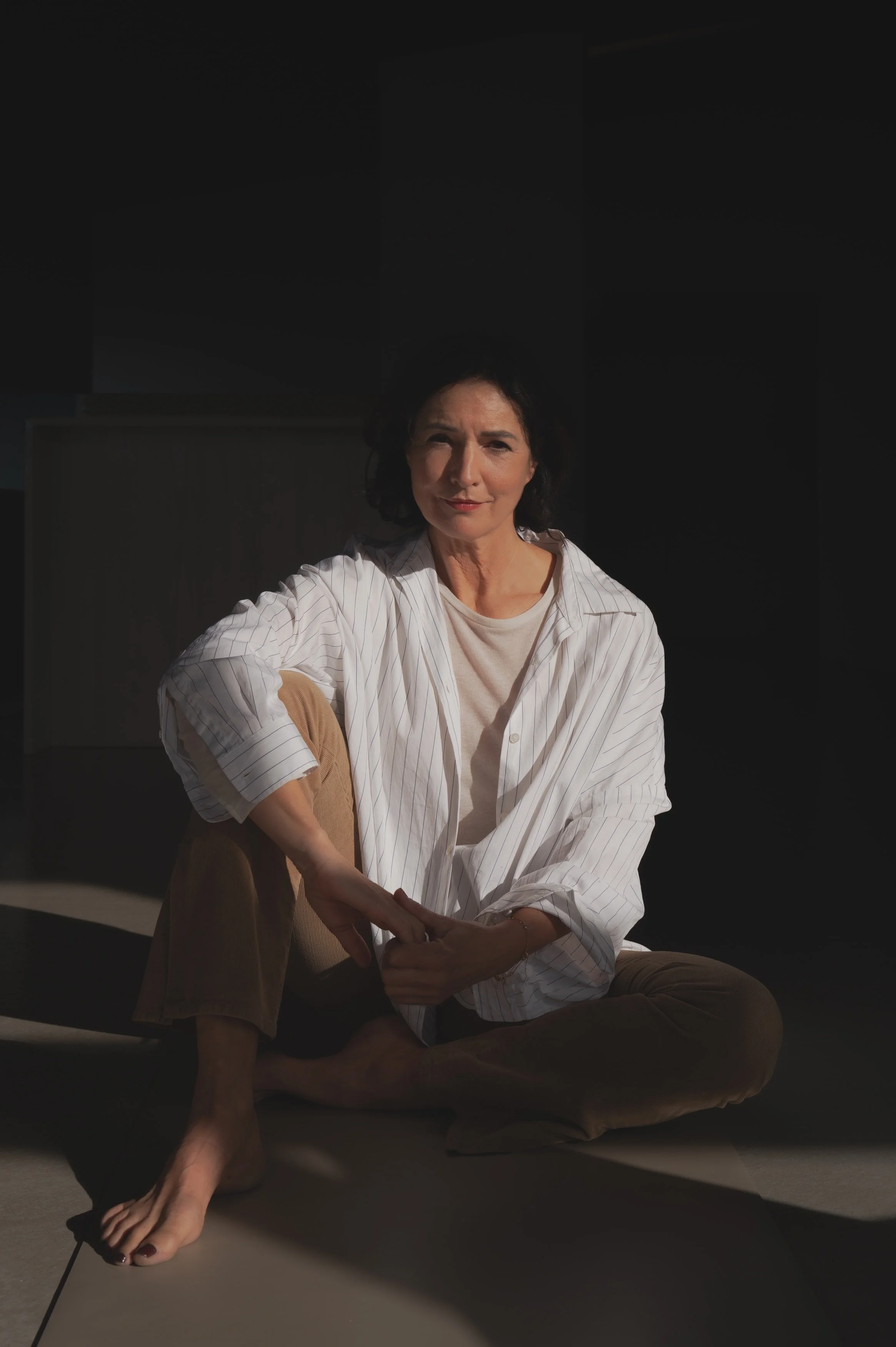 Eine Frau sitzt auf dem Boden in einem Raum, trägt ein weißes gestreiftes Hemd und braune Hosen, und schaut in die Kamera, umgeben von dunklem Hintergrund.
