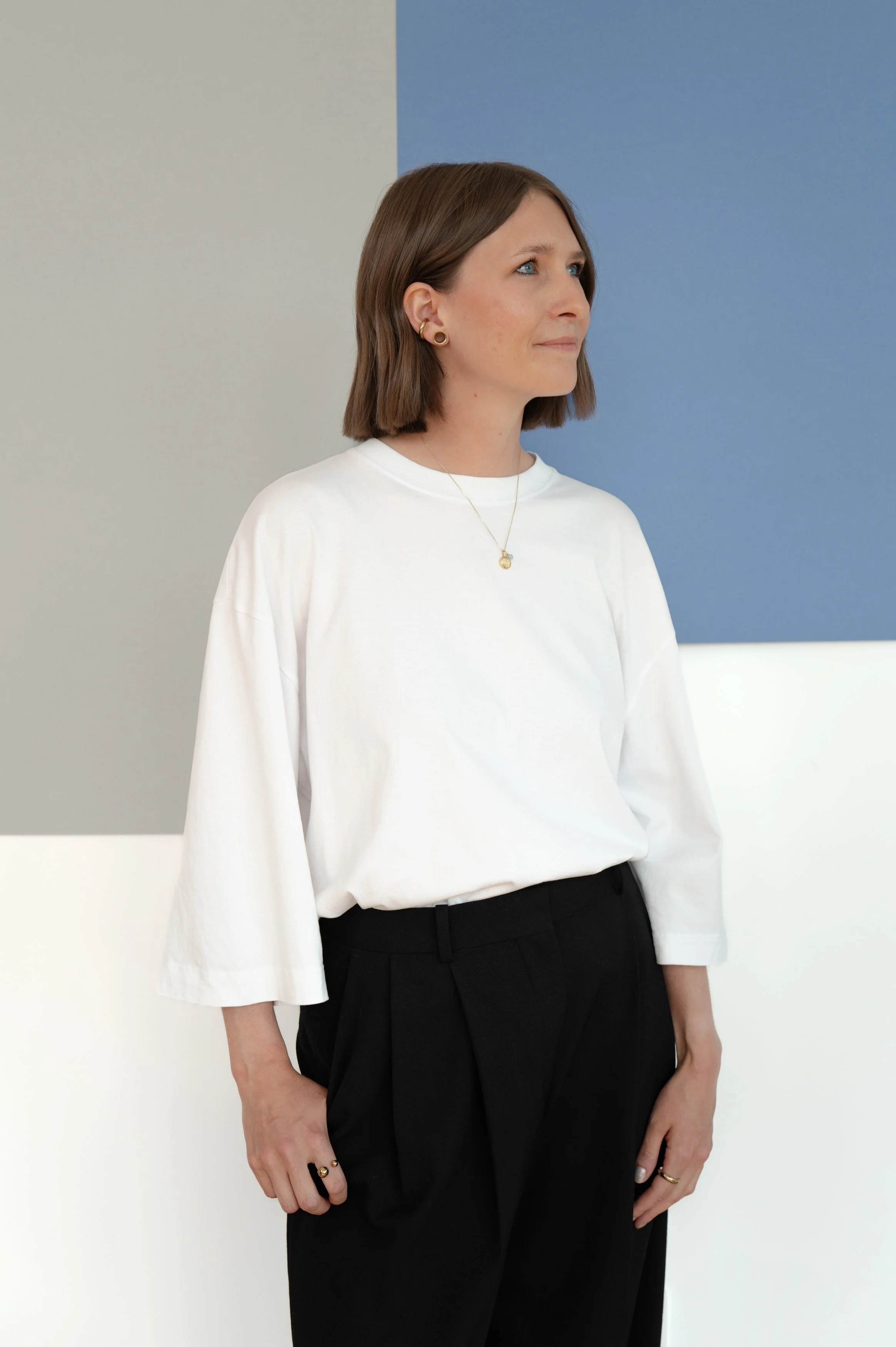 Eine junge Frau mit braunen, lockeren Haaren trägt einen weißen Pulli und schwarze Hosen, vor einer grafischen Wand mit blauen, grauen und weißen Flächen.