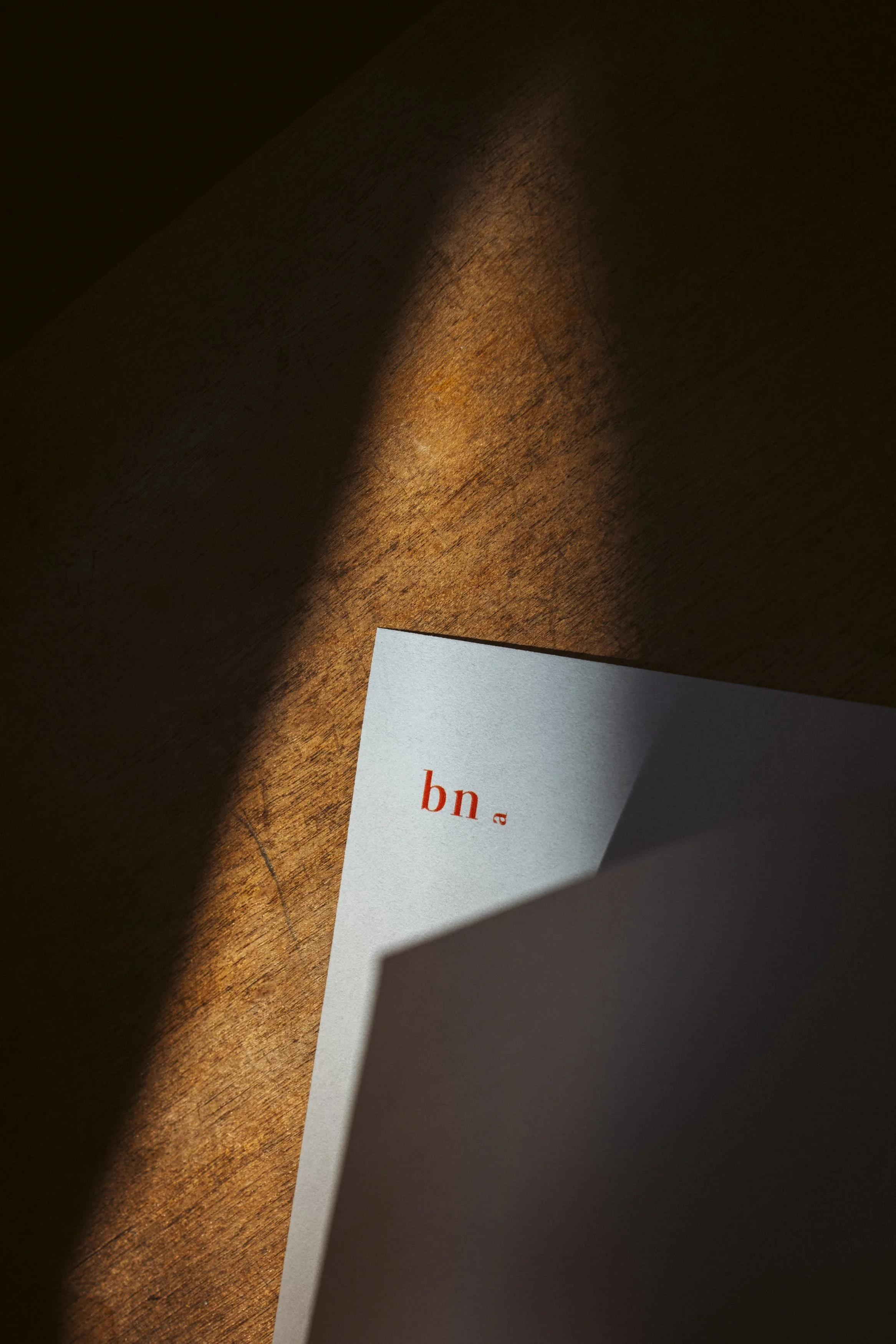 Ein Schatten fällt auf einen braunen Holz Tisch, auf dem eine weiße Karte liegt. Auf der Karte sind rote Buchstaben zu sehen, die nur teilweise erkennbar sind.