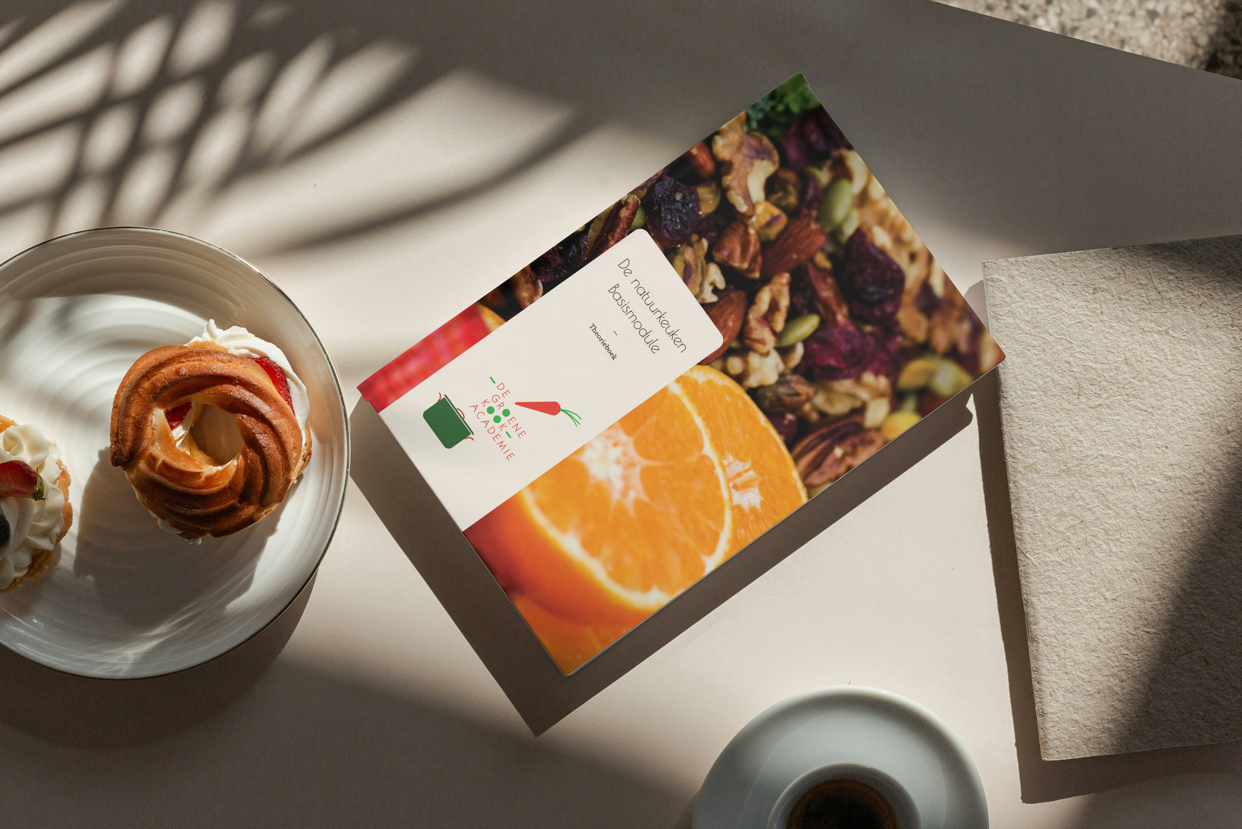 Boek op a4 formaat van bovenaf gezien met een foto van sinaasappelen en noten op de voorkant. Links ligt een bagel met cream cheese op een bordje en rechts onder zien we een koffie kopje met zwarte koffie.