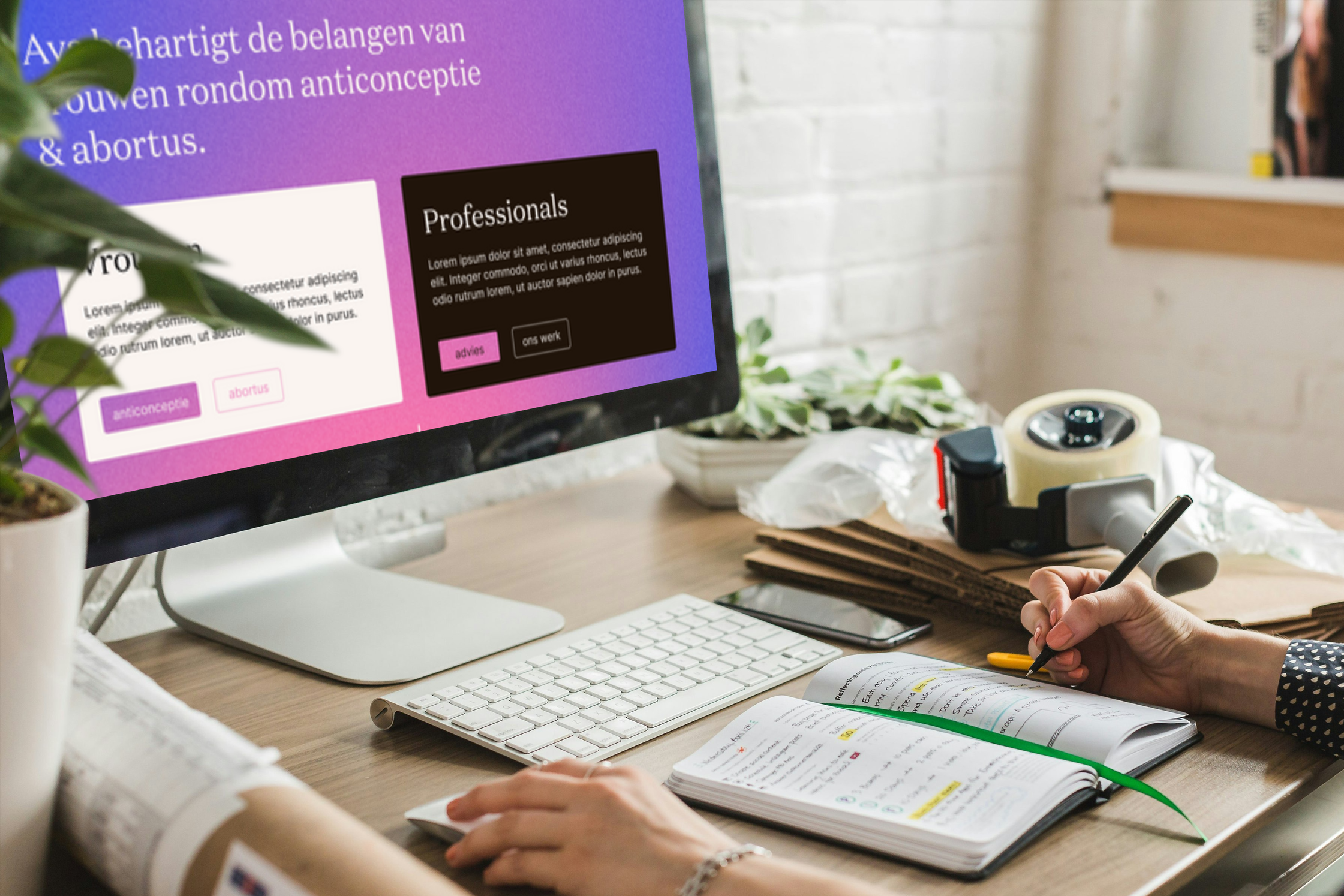 Een bureau met een computer, een telefoon, een boek en een persoon die schrijft. Op het scherm van de computer is een website zichtbaar. Er liggen planten en kartonnen dozen op het bureau.