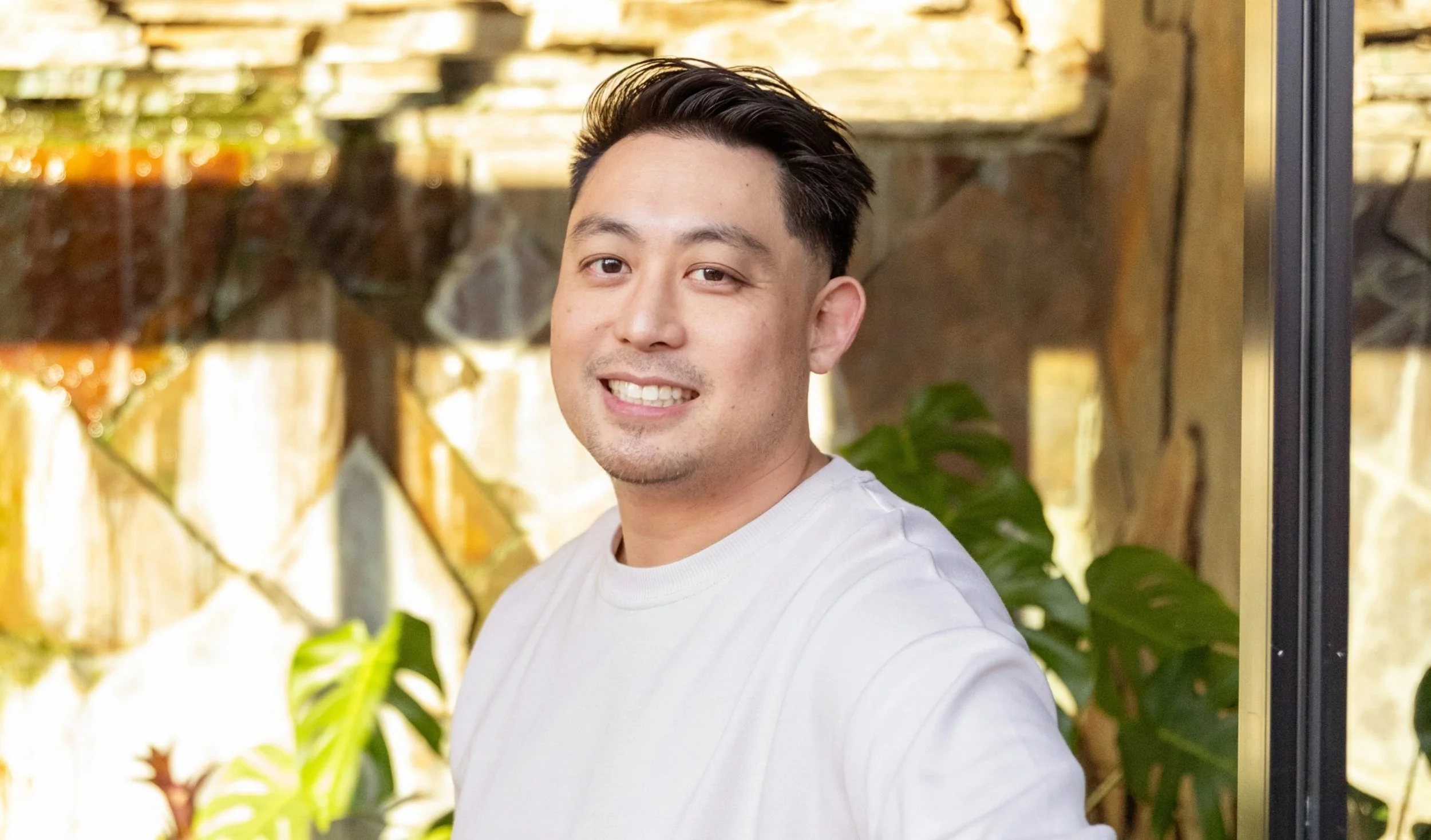 A young man taking a selfie outdoors, smiling, with a stone wall, plants, and sunlight in the background.