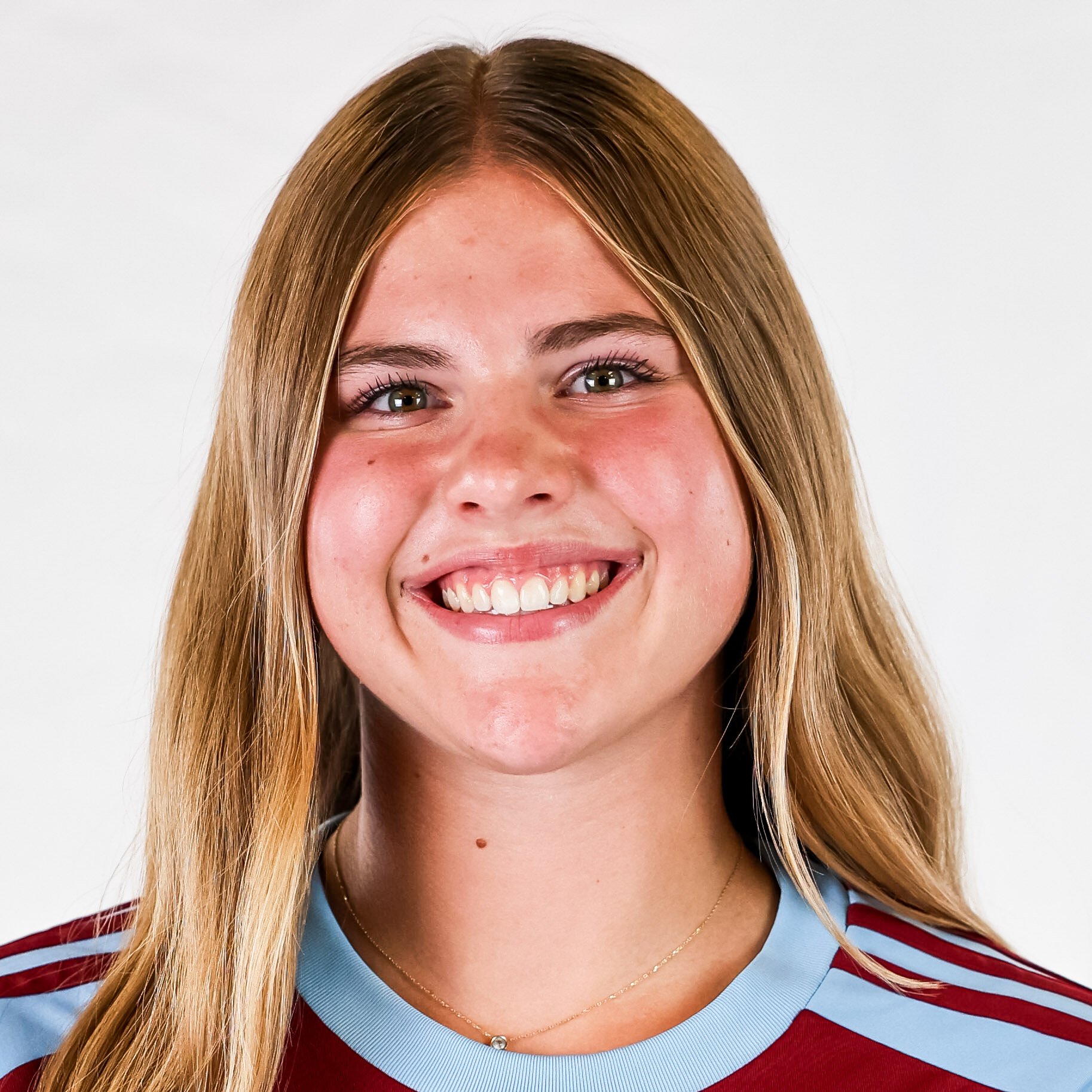 A young woman with long blonde hair smiling at the camera, wearing a maroon and light blue sports jersey, and a delicate necklace.