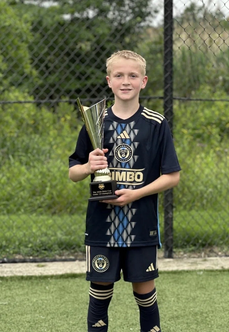 A young boy in a soccer uniform holding a trophy, standing on a grassy field with a chain-link fence and green trees in the background.