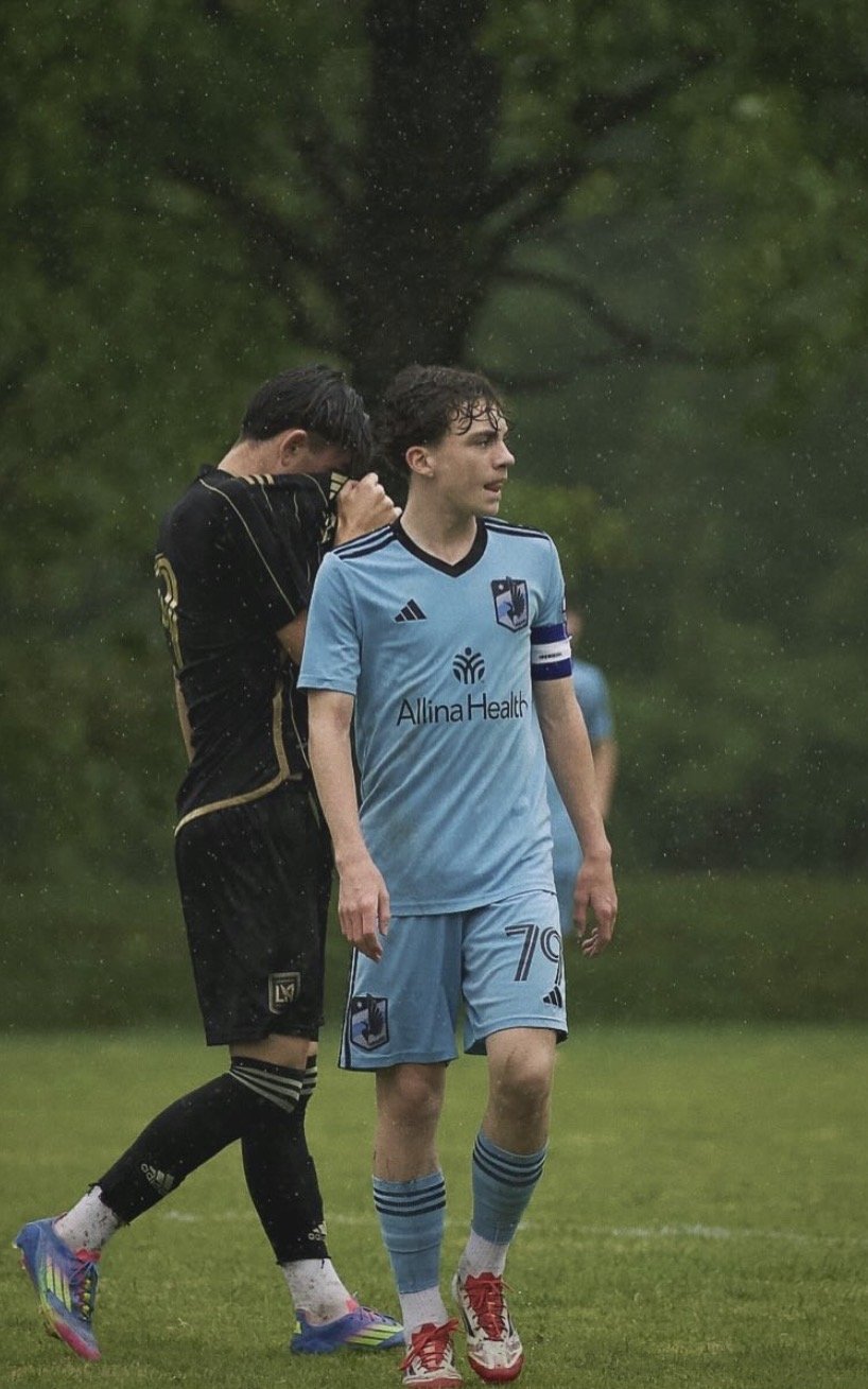 Two soccer players walking on a rainy field, one in a black uniform and the other in a light blue uniform, with trees and rain in the background.
