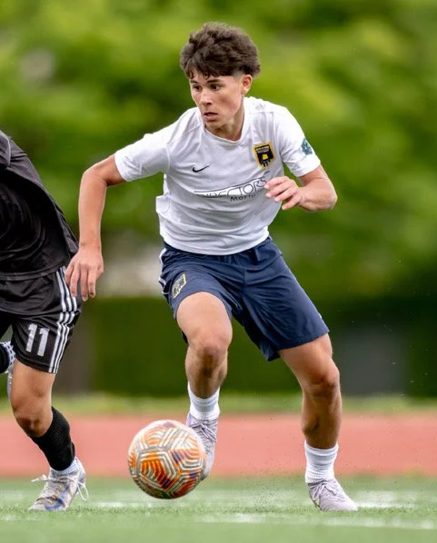 A young male soccer player in a white jersey and navy shorts dribbling a soccer ball during a game.
