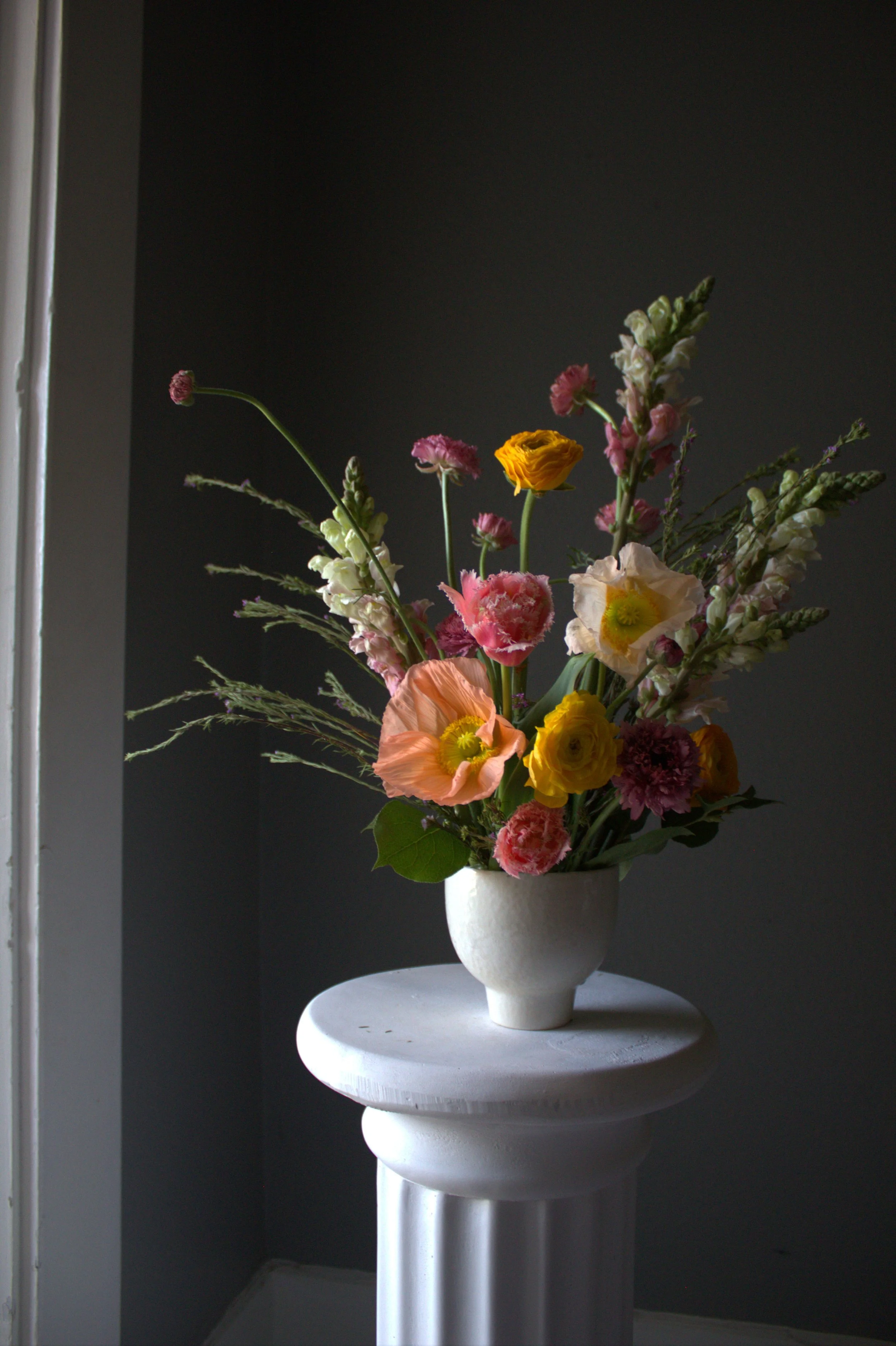 Sculptural floral arrangement featuring peach poppies, yellow ranunculus, and pink seasonal flowers in a white vase for Dahlias & Daisies Bloom Club.