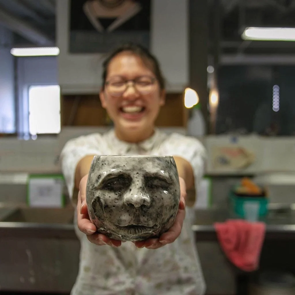 A clay cup in the shape of the moon with a face on it and the student who made it smiling while they hold it