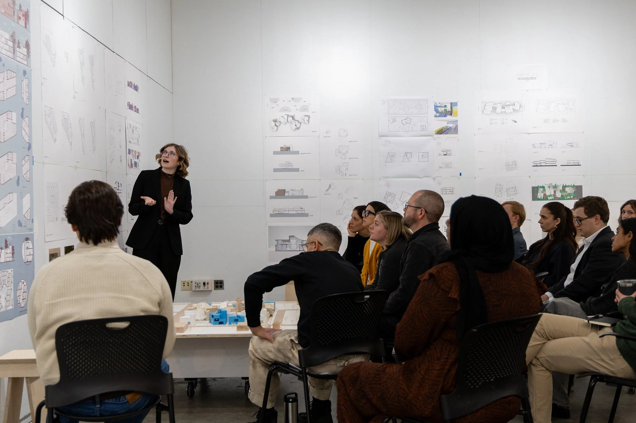 A student dressed in black presents their work to a group of faculty and peers