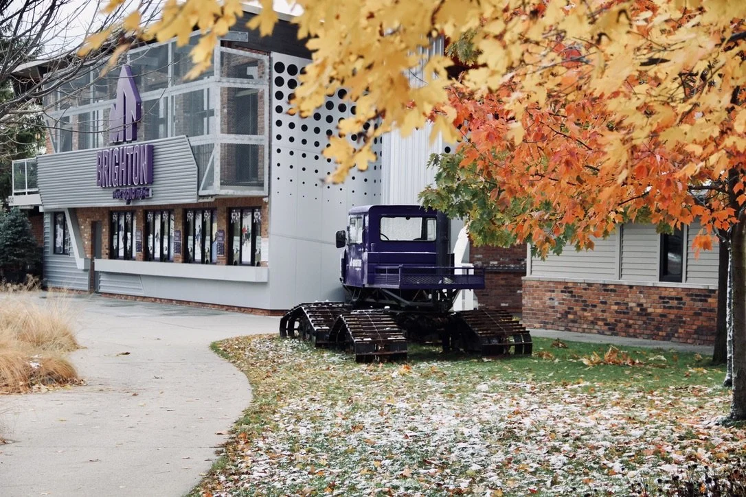 A vintage snowcat parked under a fall colored tree and next to the entrance of a ski resort