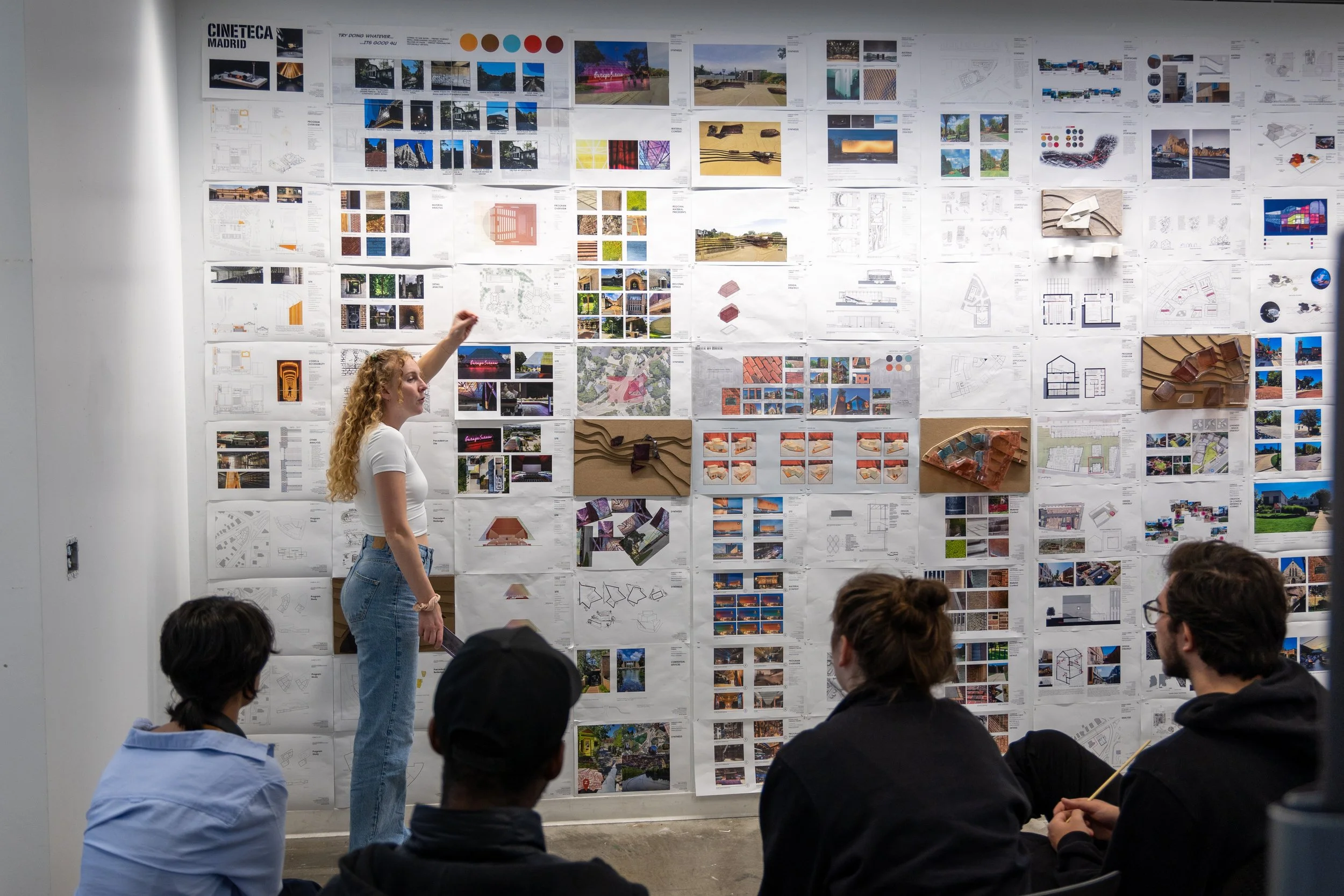 A student presents their work pinned up on a large wall in front of a panel of judges