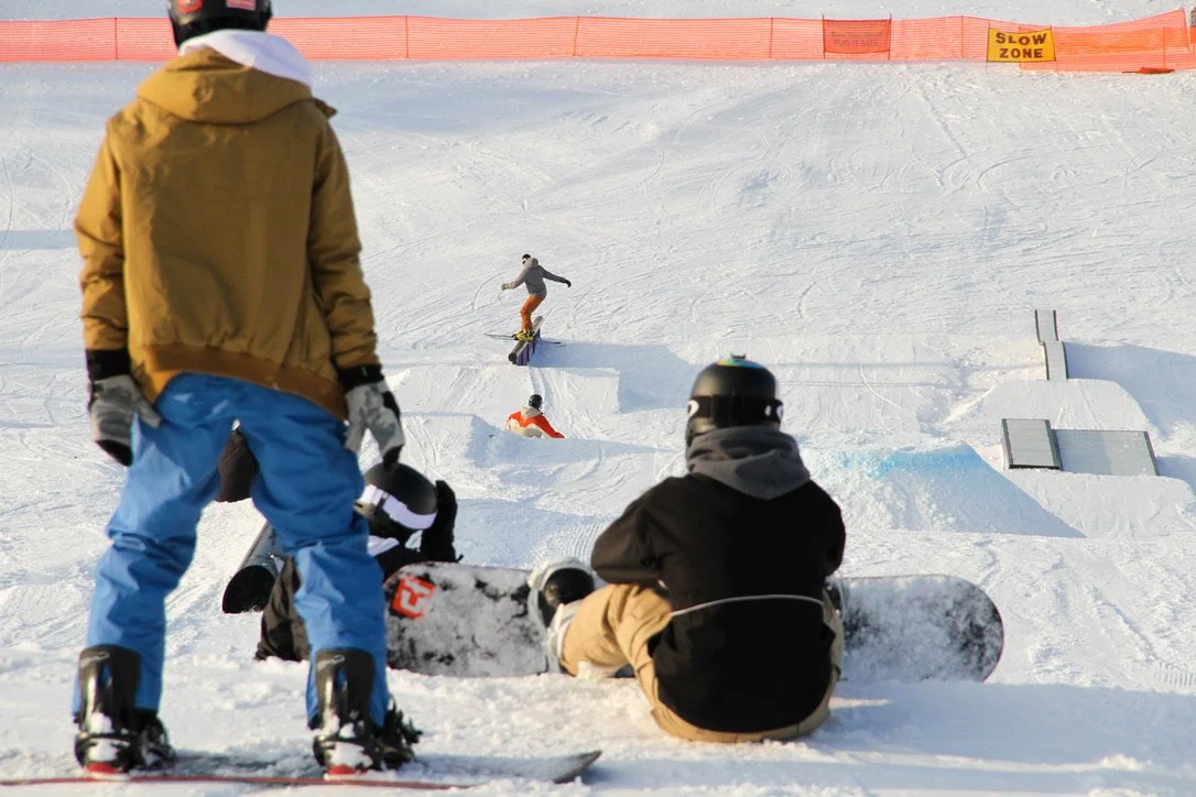 Snowboarders sit at the top of the train park watching other snowboarders on the features