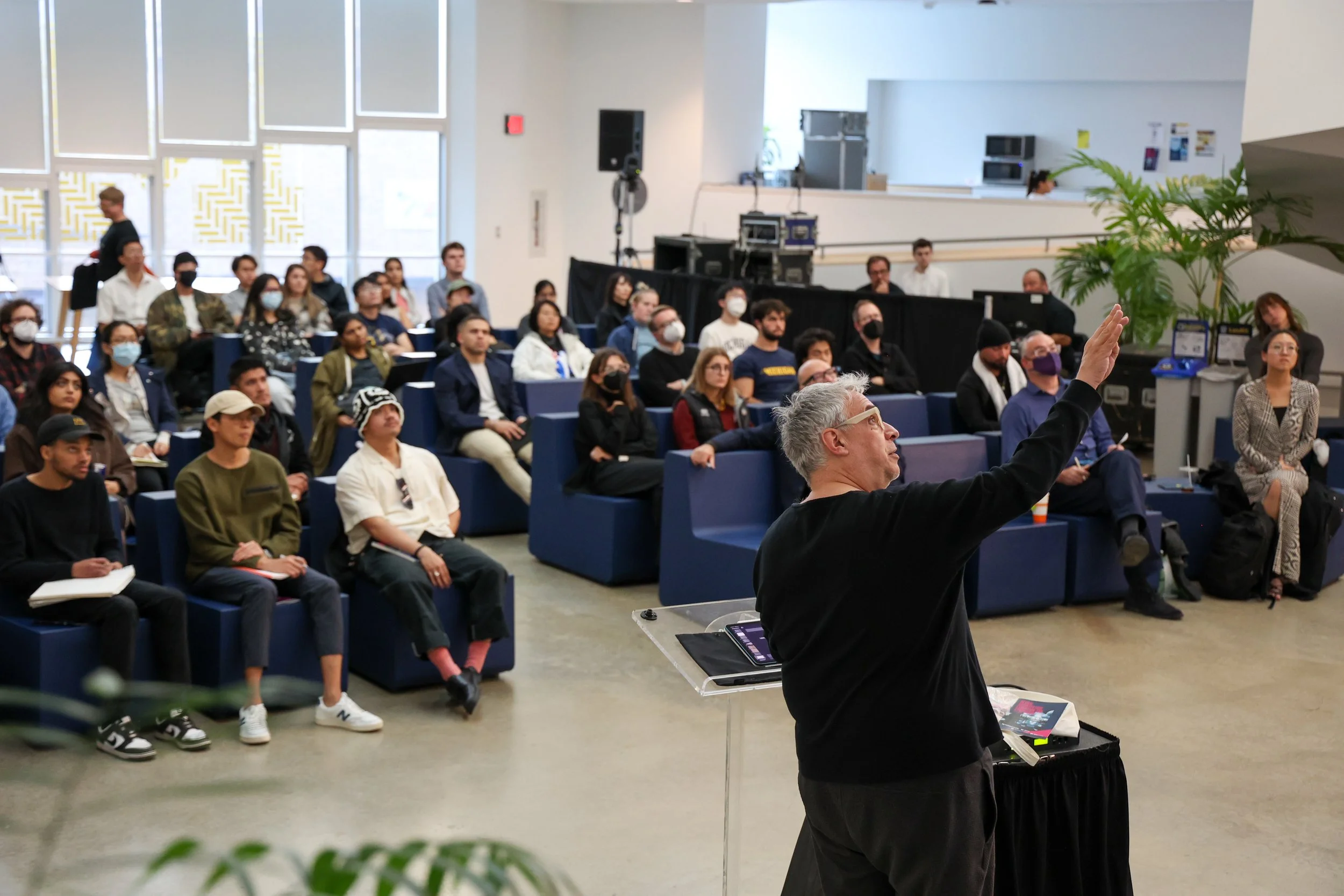 A man gives a lecture pointing up to a screen with a crowd of people seated in front of him