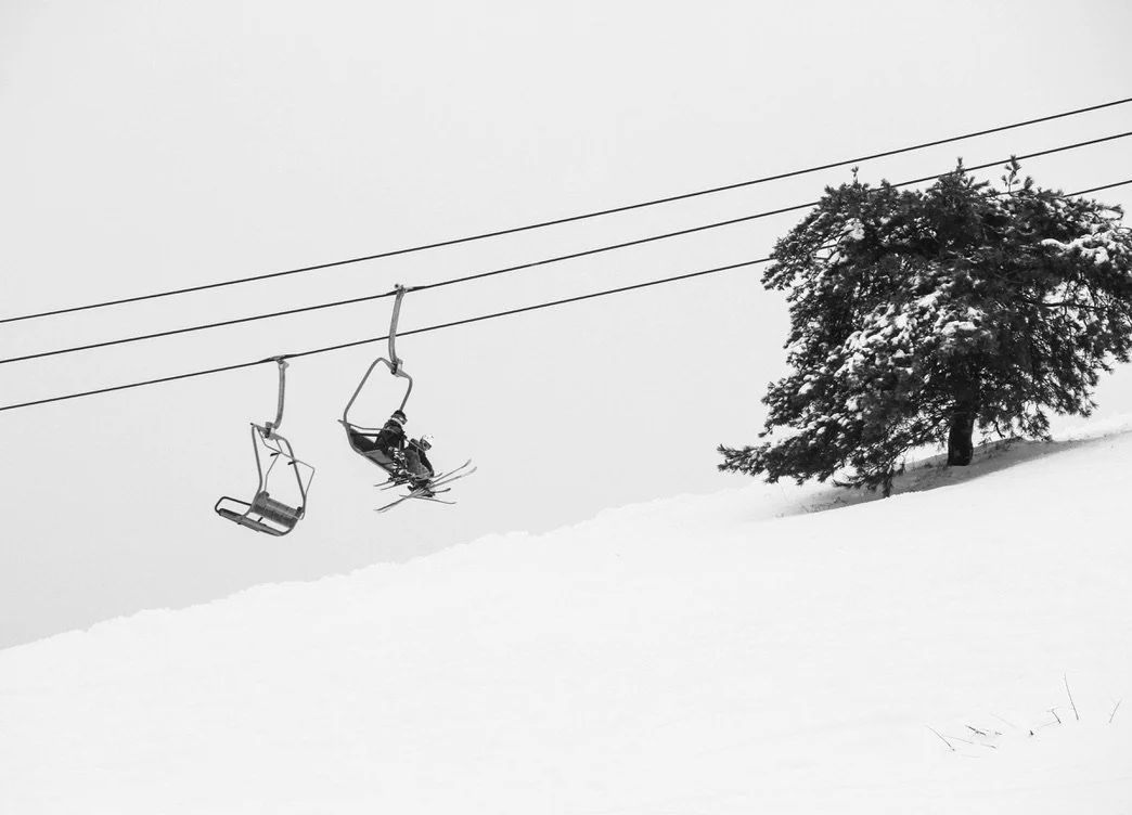 Three skiers ride up a chair lift with a pine tree and snow in the foreground