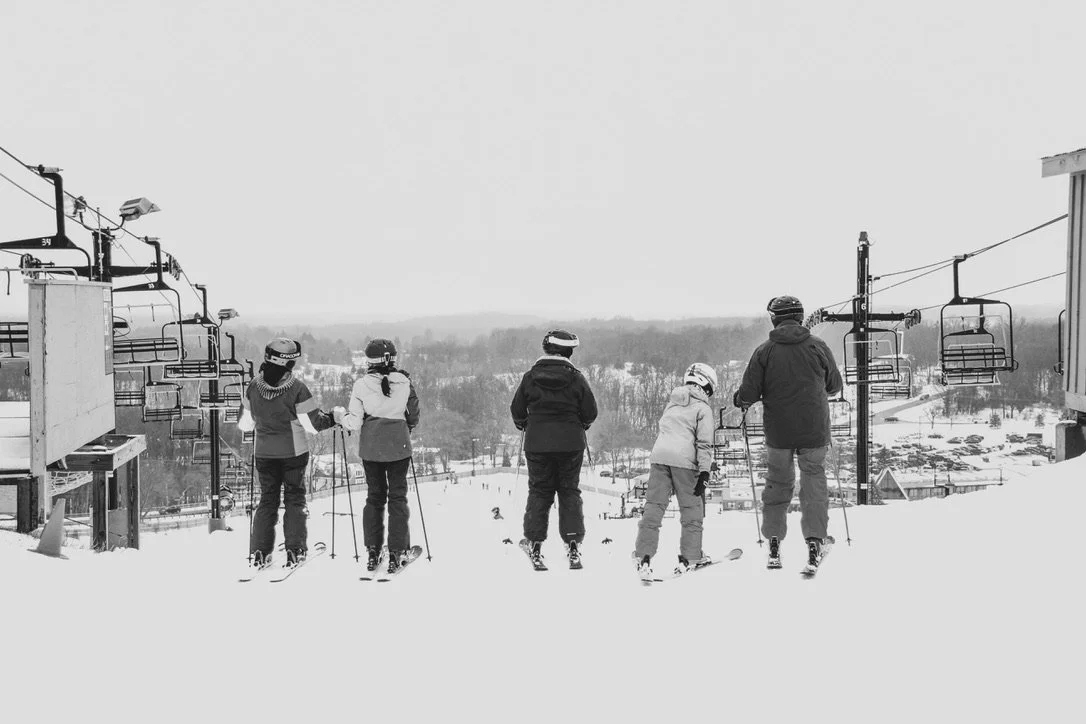 A row of five people about to descend from the top of a ski resort mountain