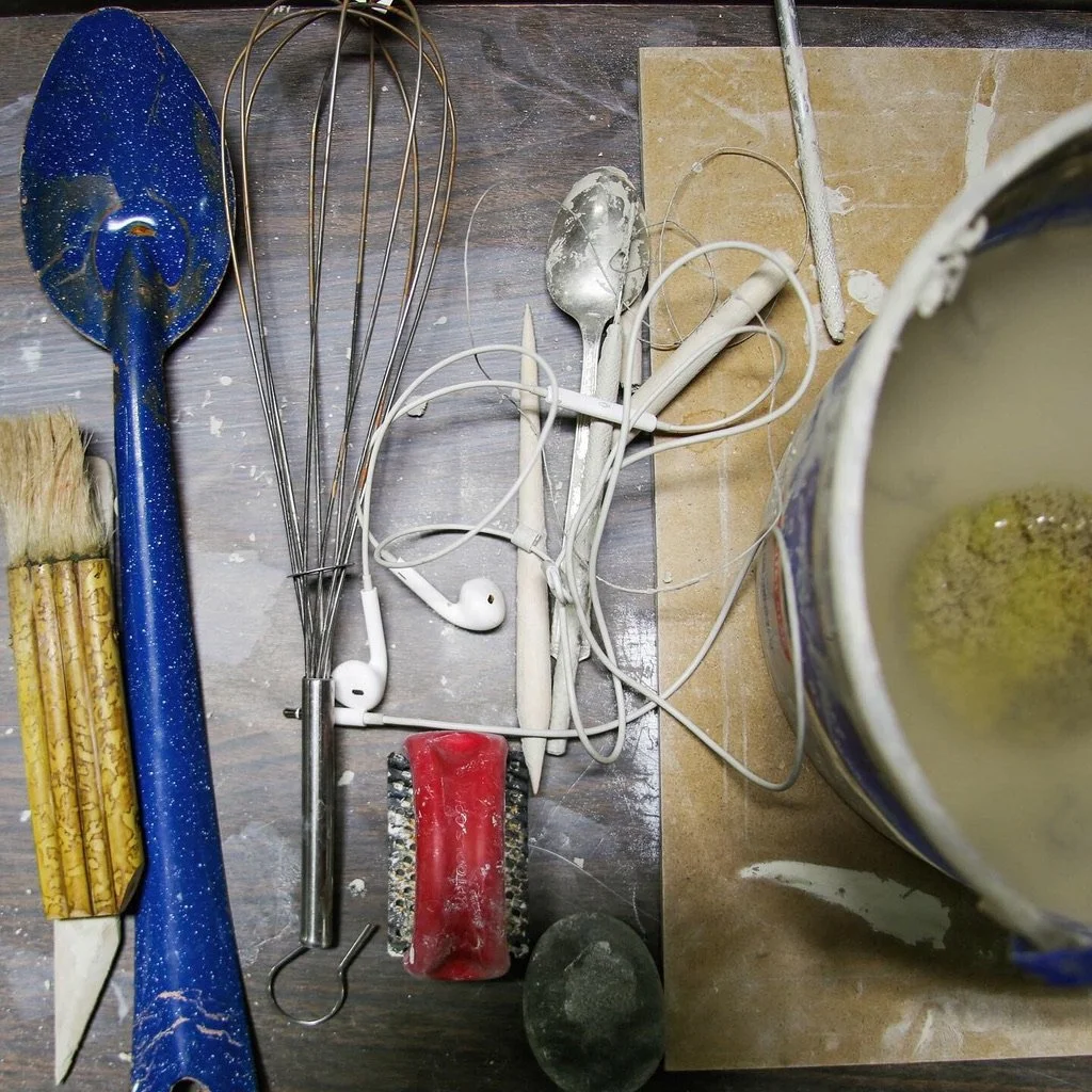 A top down view of a pottery students desk featuring s spoon, whisk, headphones, bucket and other tools
