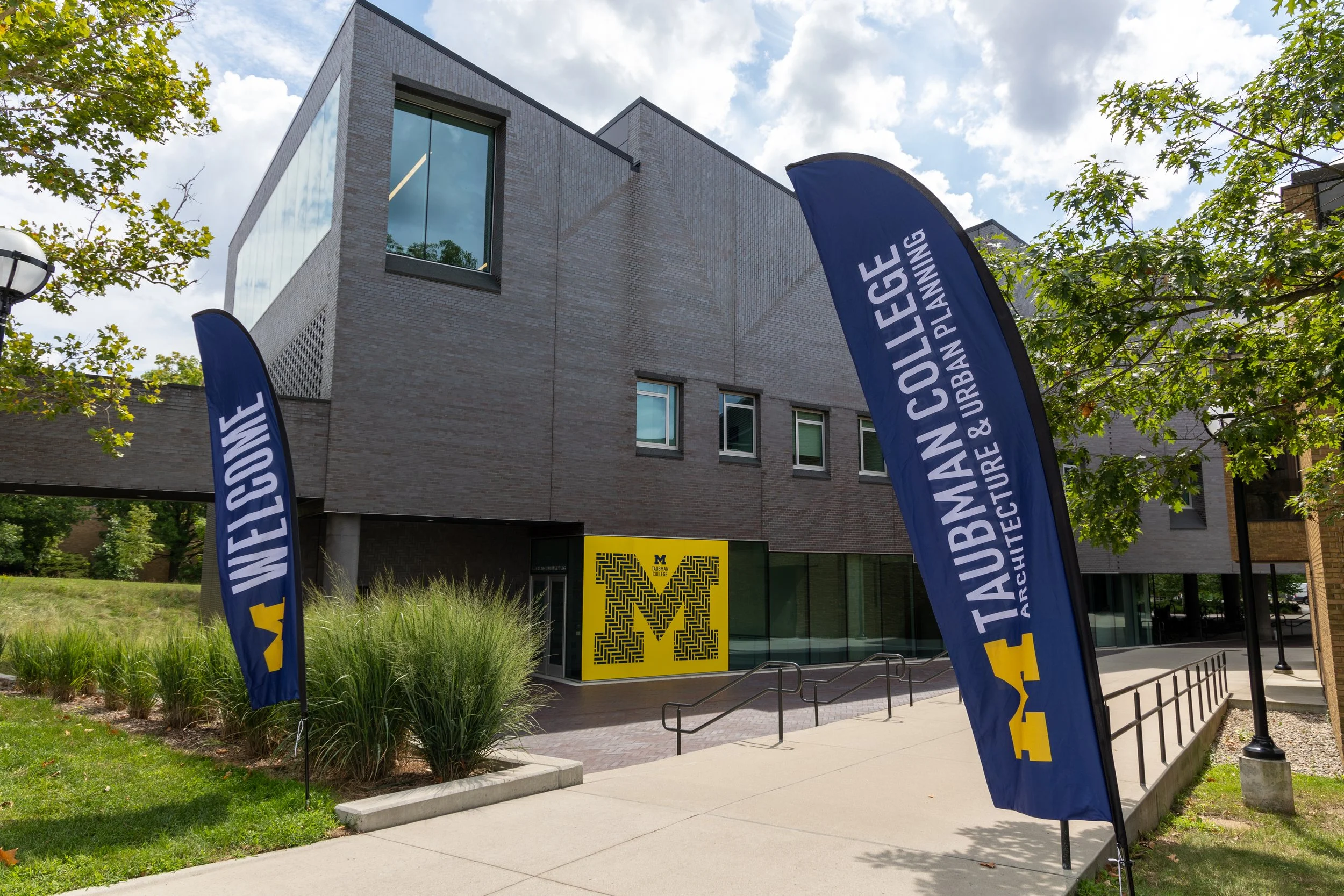 The exterior of a building at the University of Michigan. Two welcome flags wave with a big yellow block M on one of the glass panes