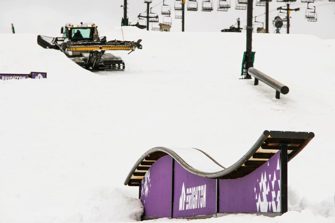 A snowcat climbs up hill in the background and a terrain park feature is in focus in the front