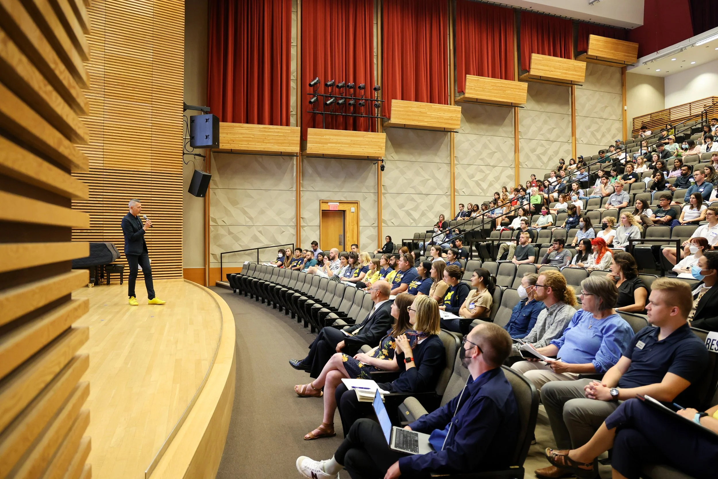a speaker standing on stage talking to a large crowd in an auditorium 
