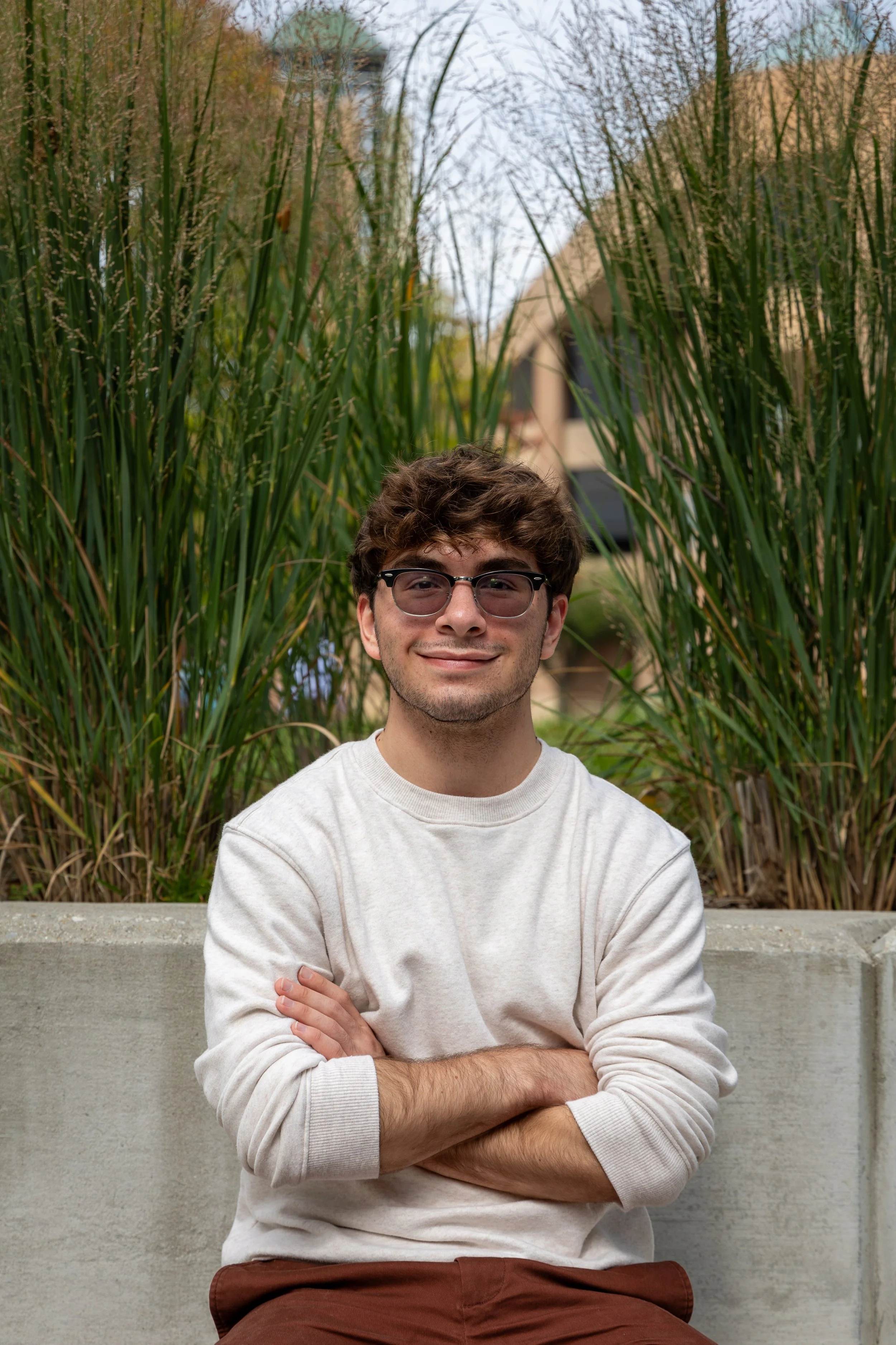 A headshot of a man with his arms crossed, sitting on a concrete bench with tall grasses behind him