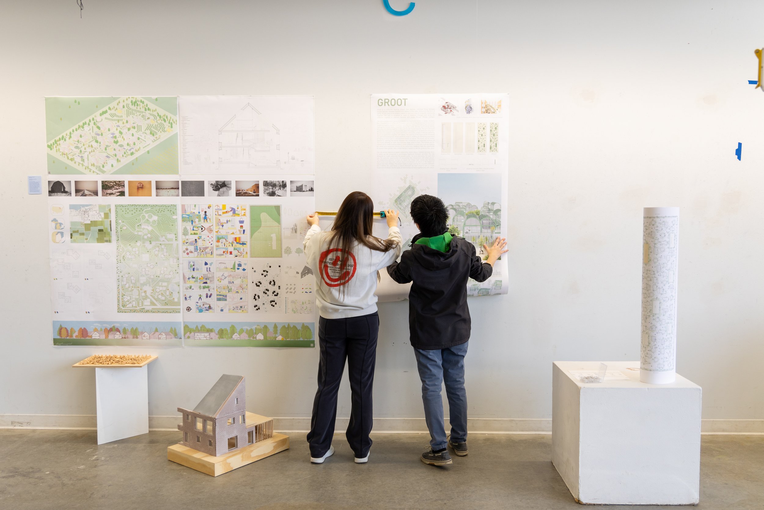 Two students facing a wall meaure their work that is being pinned up