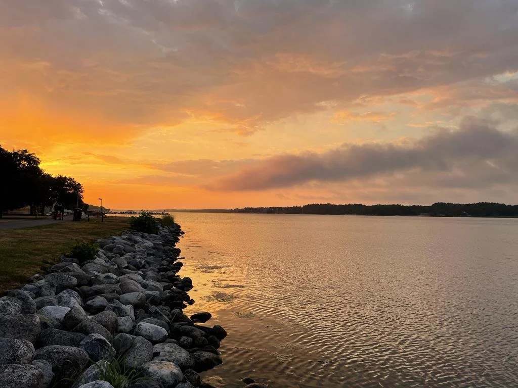 En lugn sjö vid solnedgång, med en stenstrand och träd längs kanten, molnigt himmel med orange och gula nyanser.