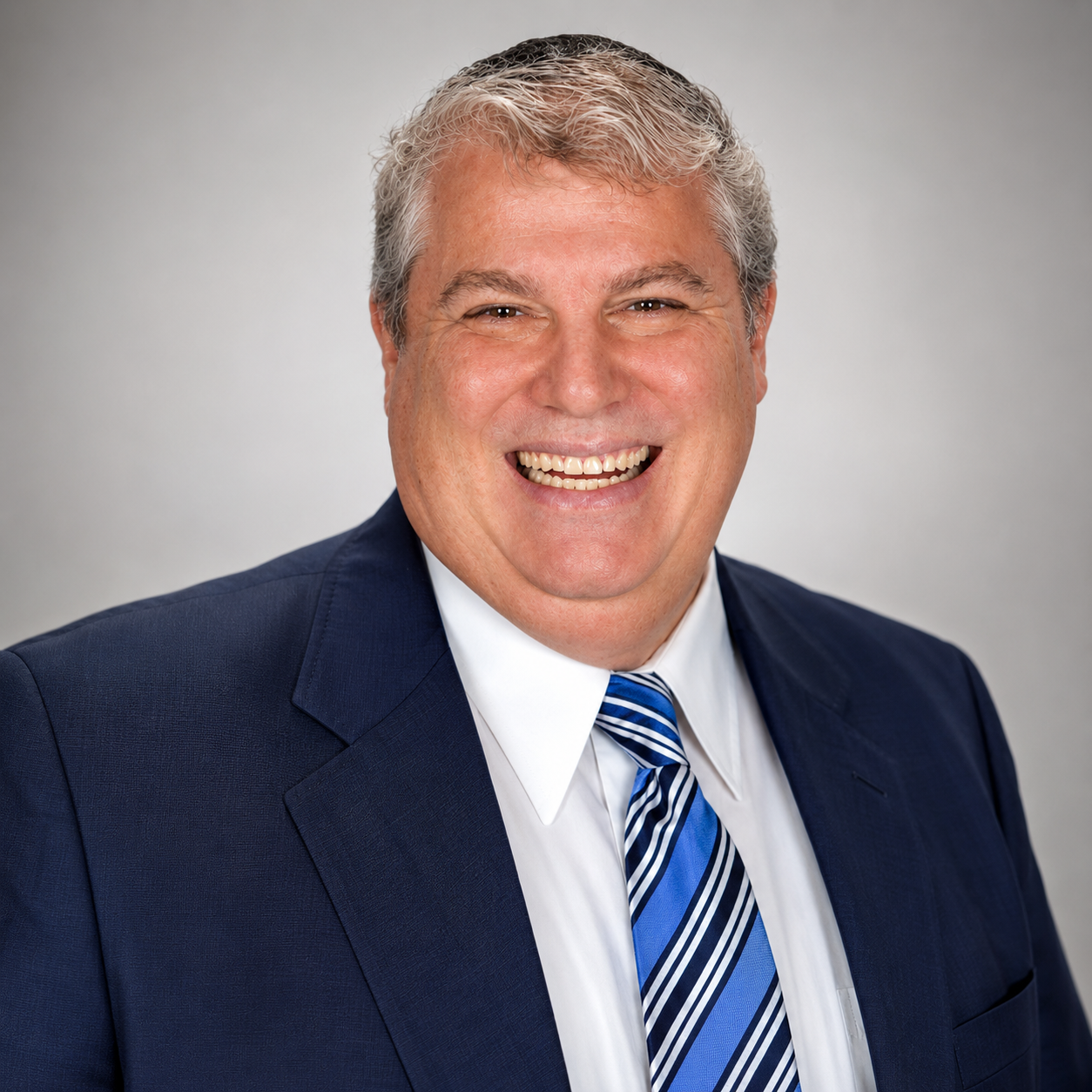 A smiling middle-aged man with gray hair, wearing a dark suit, white shirt, and a blue striped tie, standing against a plain white background.