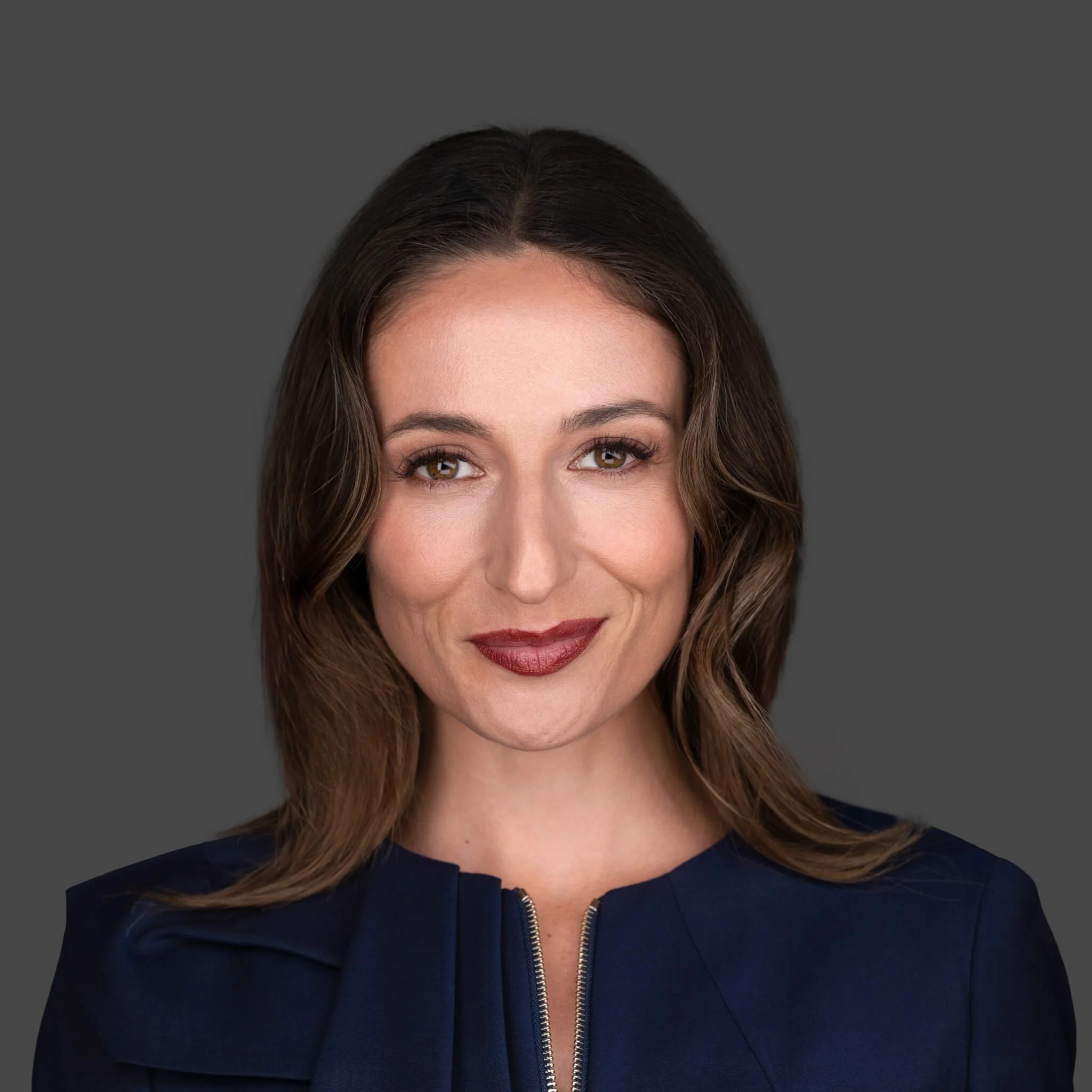 A woman with dark hair in a short hairstyle, wearing a black turtleneck and black embroidered jacket, standing against a gray background.