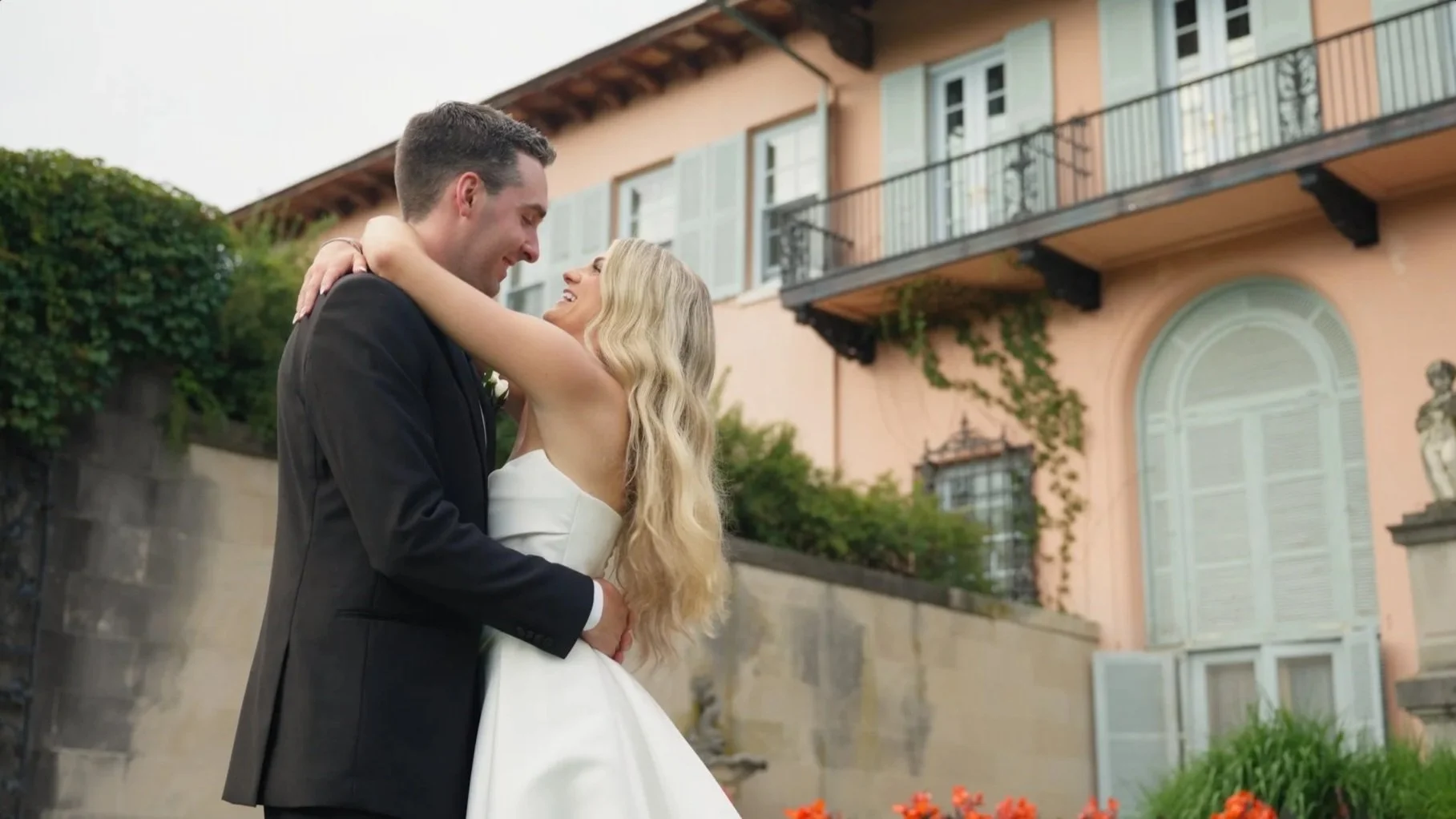 A bride and groom dancing outdoors in front of a pink house with green shutters, with greenery and orange flowers in the foreground.