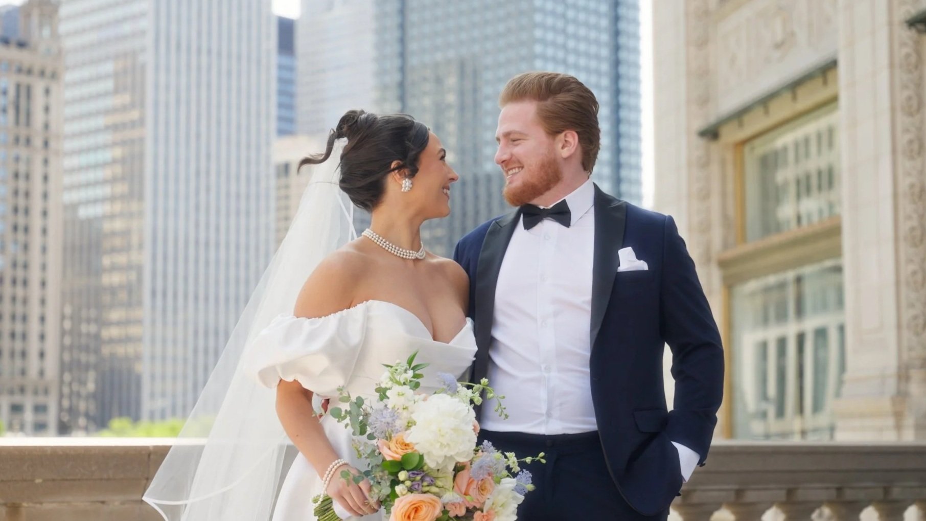 A newlywed couple in wedding attire smiling at each other outdoors in an urban city setting with tall buildings in the background.