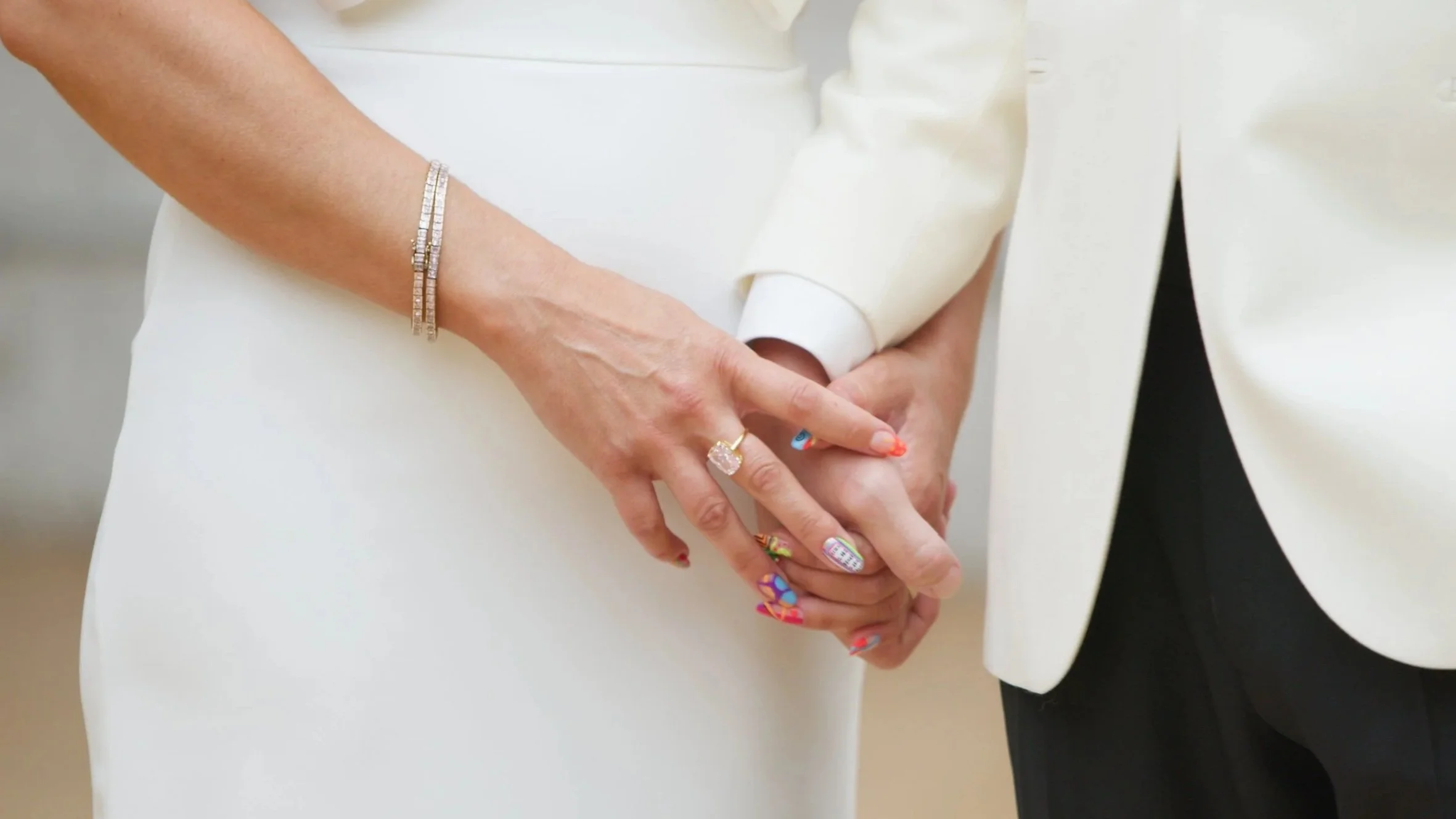 A man and woman holding hands, dressed in elegant clothing. The woman has a diamond ring and colorful, decorated nails, and is wearing a diamond bracelet. The man is dressed in a white suit with a black shirt underneath.