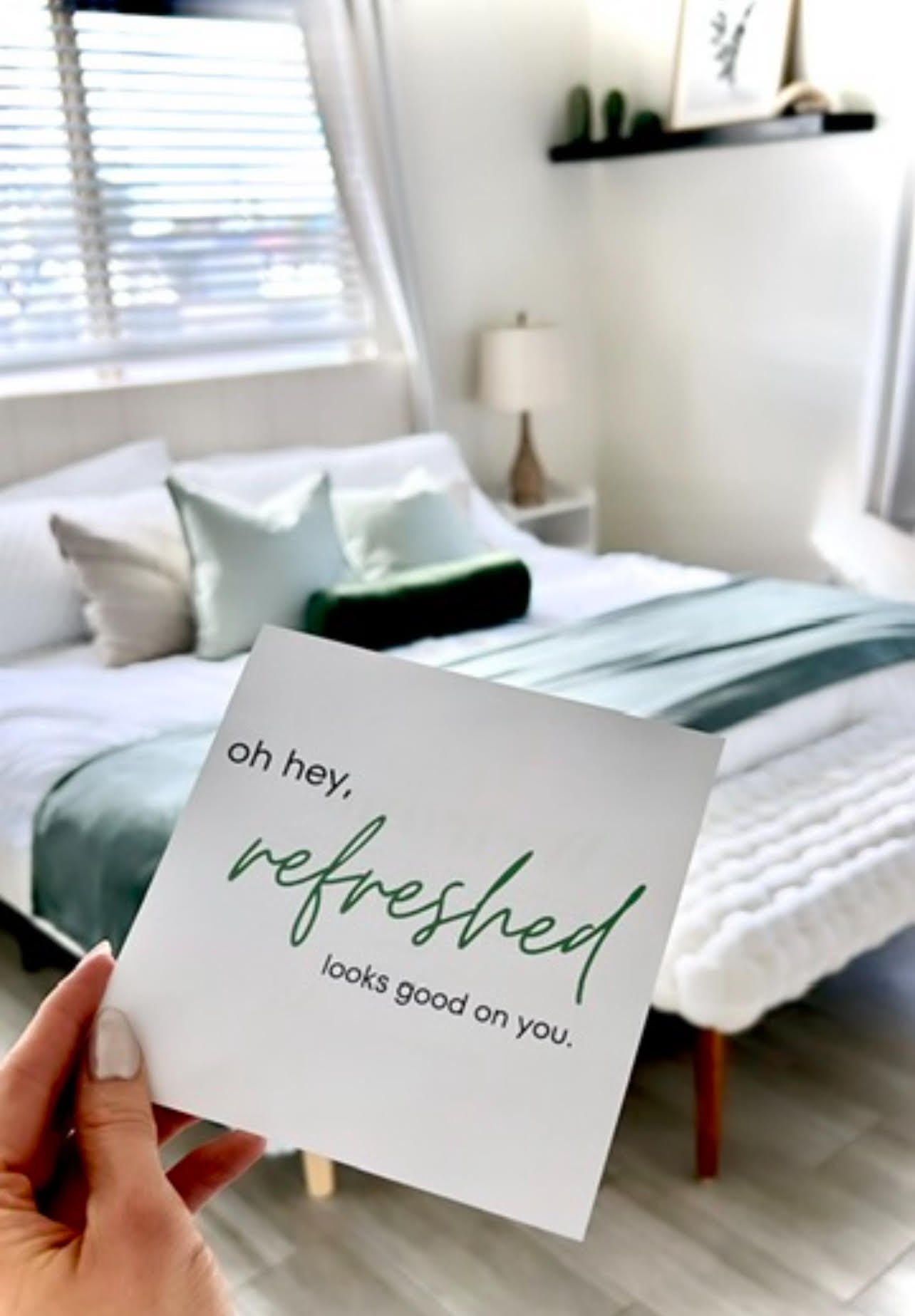 A person holding a white card with the text "oh hey, refreshed looks good on you." in front of a neatly made bed with white pillows, a gray and white striped blanket, a bedside table with a lamp, and a window with blinds in a bright, modern bedroom.