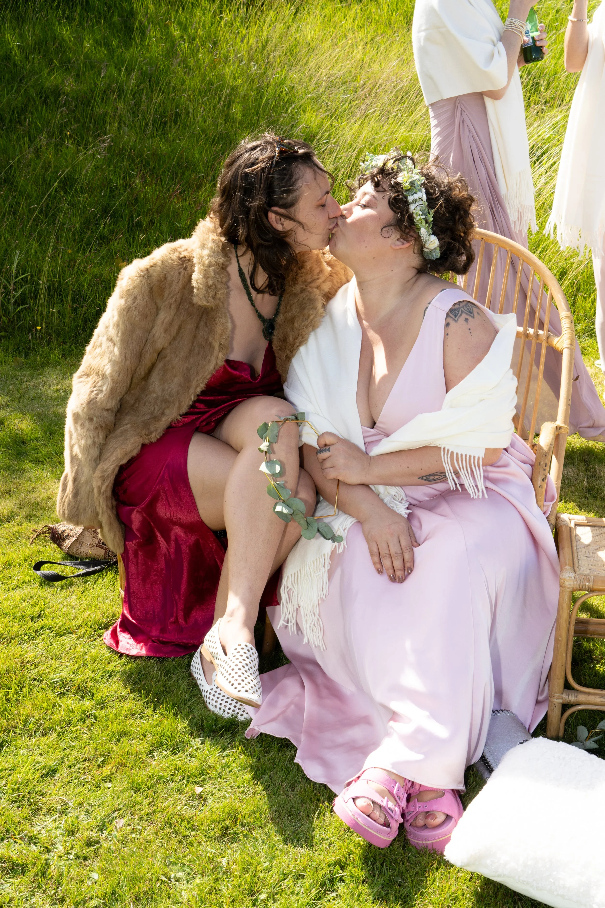 Two women are sitting on a chair outdoors, sharing a kiss during a celebration or gathering on a grassy area, with other people standing in the background.