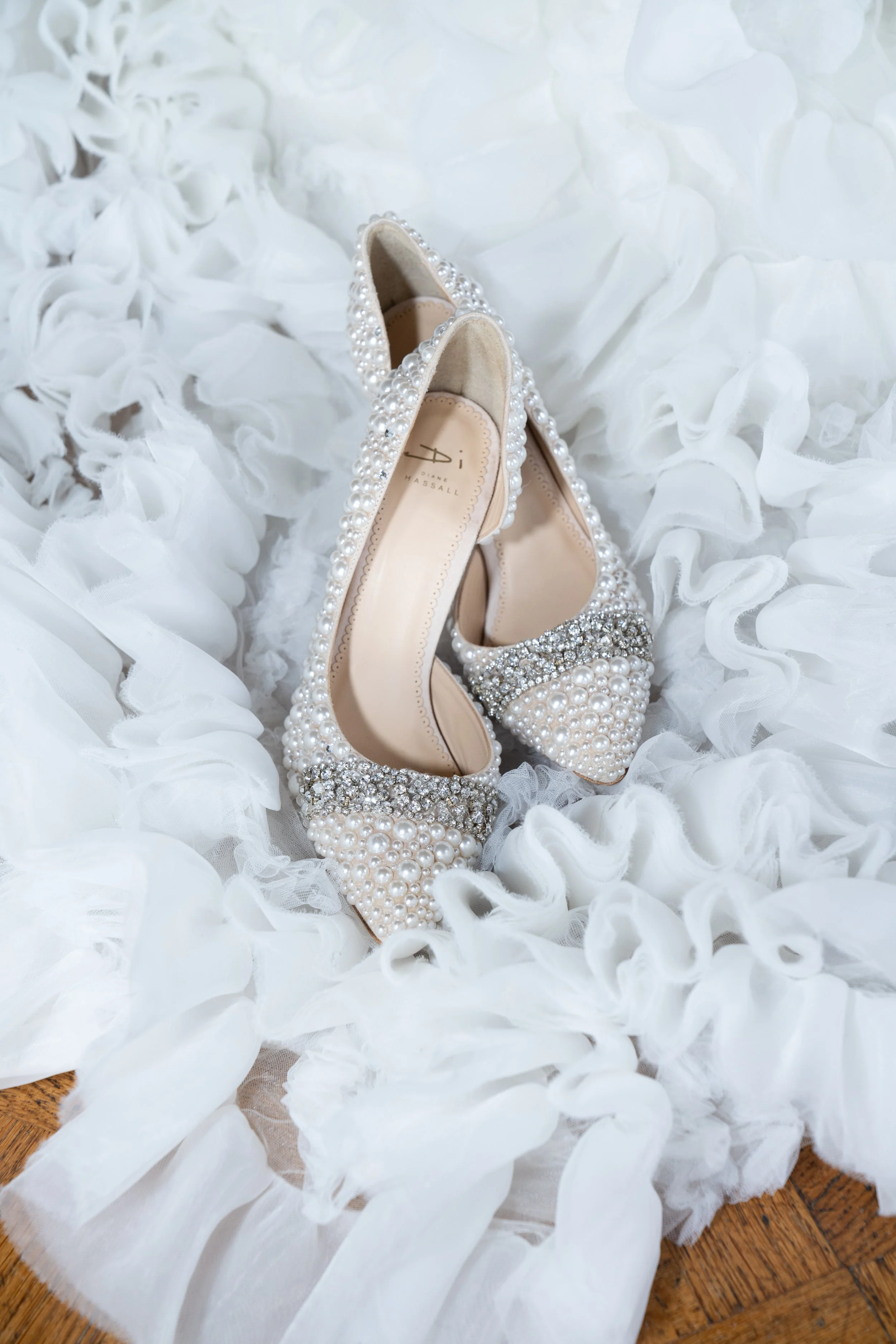A pair of white high-heeled shoes for a wedding decorated with pearls and rhinestones, placed on white ruffled dress.